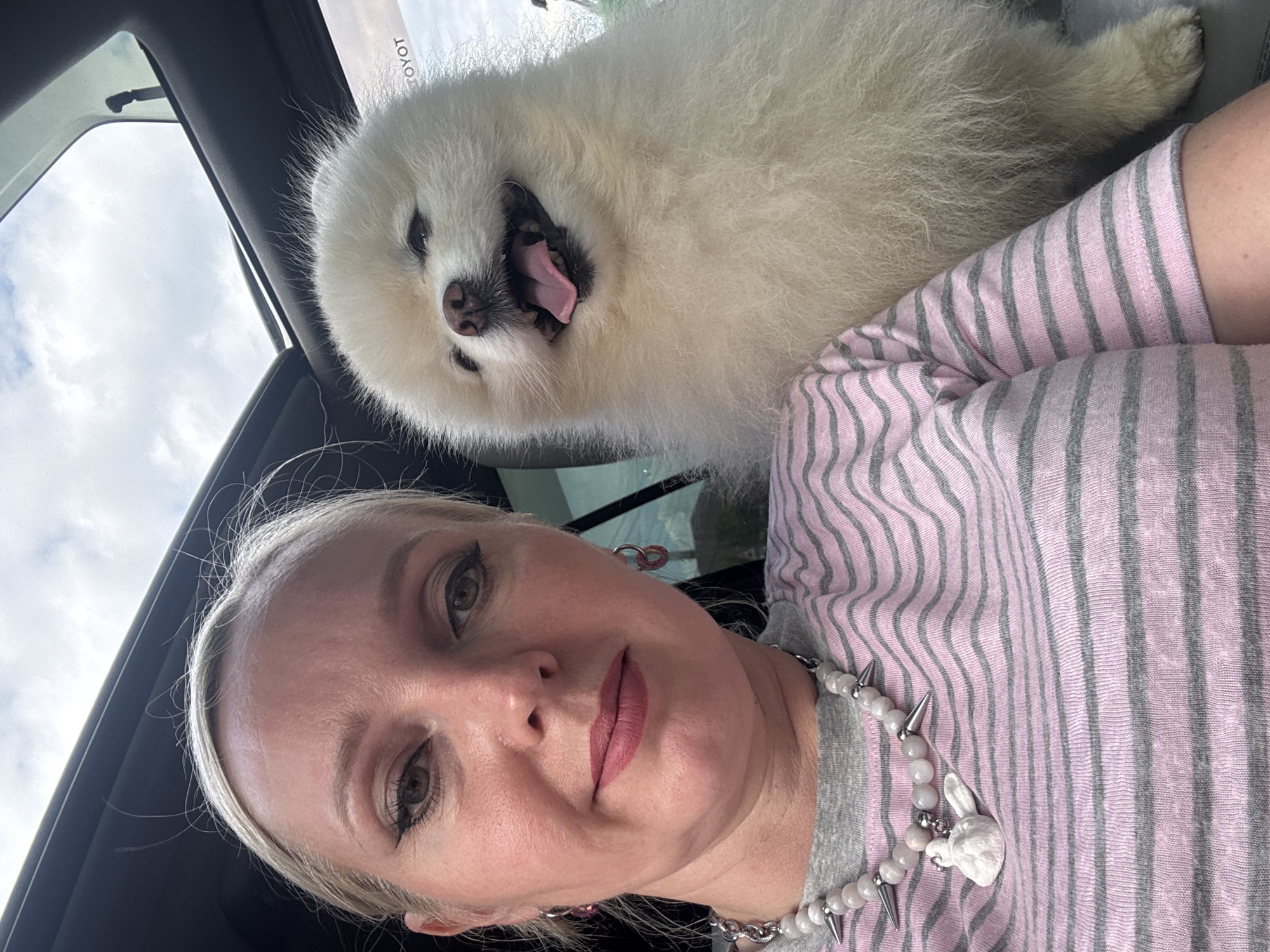 In Komsomolsk-on-Amur, Russia, a woman and a white Pomeranian dog are photographed inside a vehicle. The woman, with blonde hair and makeup including eyeliner and lipstick, is positioned on the left side of the frame, looking directly at the camera. She is wearing a pink and gray striped t-shirt and a beaded necklace with spike accents and a white bunny pendant. The Pomeranian, with its mouth open and tongue visible, is on the right side of the frame, appearing to the right and slightly behind the woman. The vehicle's sunroof is open, revealing a cloudy sky. The interior of the car, including the dashboard and part of the passenger seat, is visible.