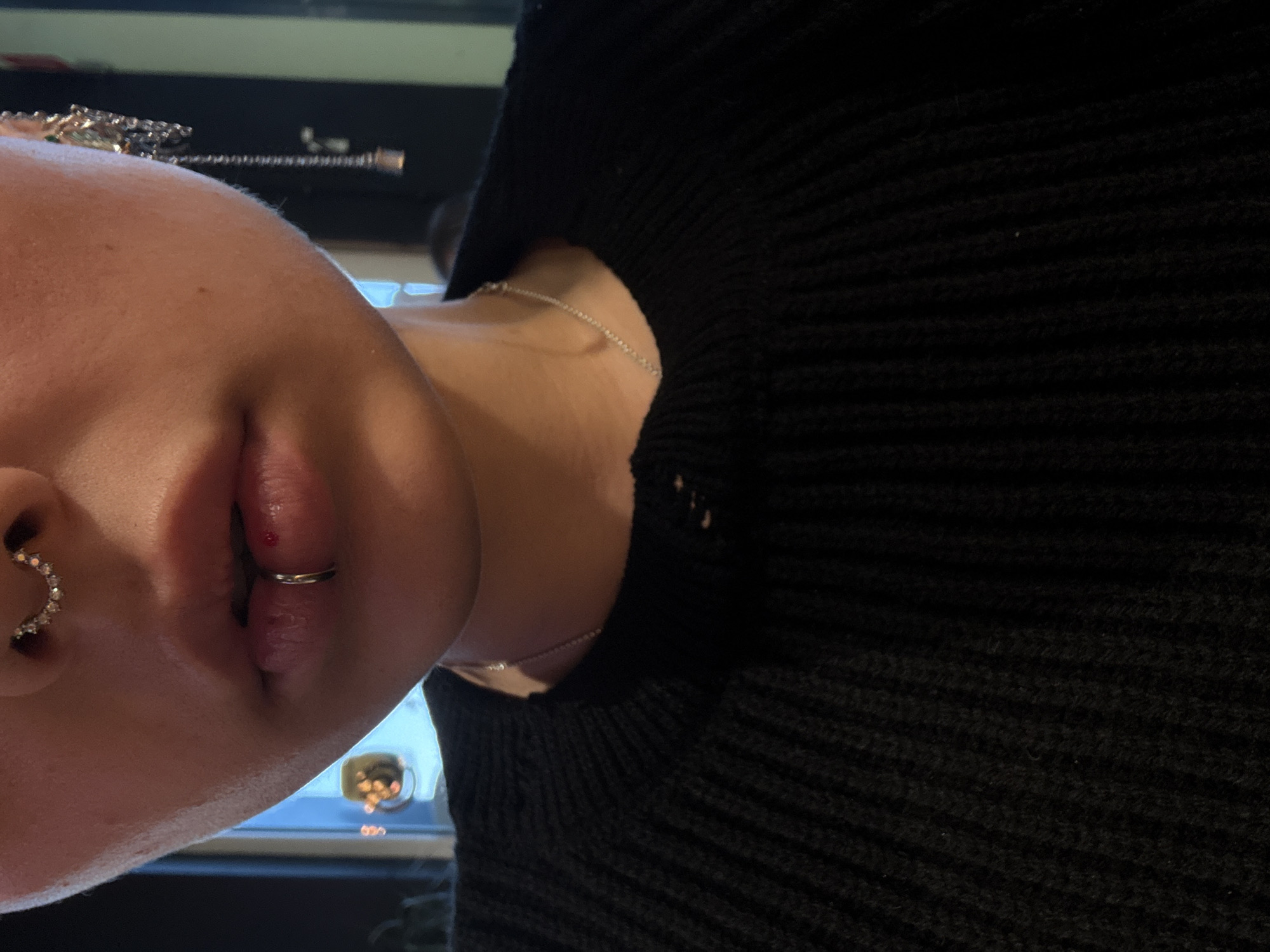 A close-up photograph depicts a person with multiple facial piercings. A ring piercing adorns their lower lip, with a small droplet of blood visible near the piercing. A crescent-shaped piercing with small gemstones is present in their nostril. The person is wearing a black knit sweater. A silver necklace is visible around their neck. The background is blurry but appears to show a window with some lights. The location cannot be determined based on the visual information provided.