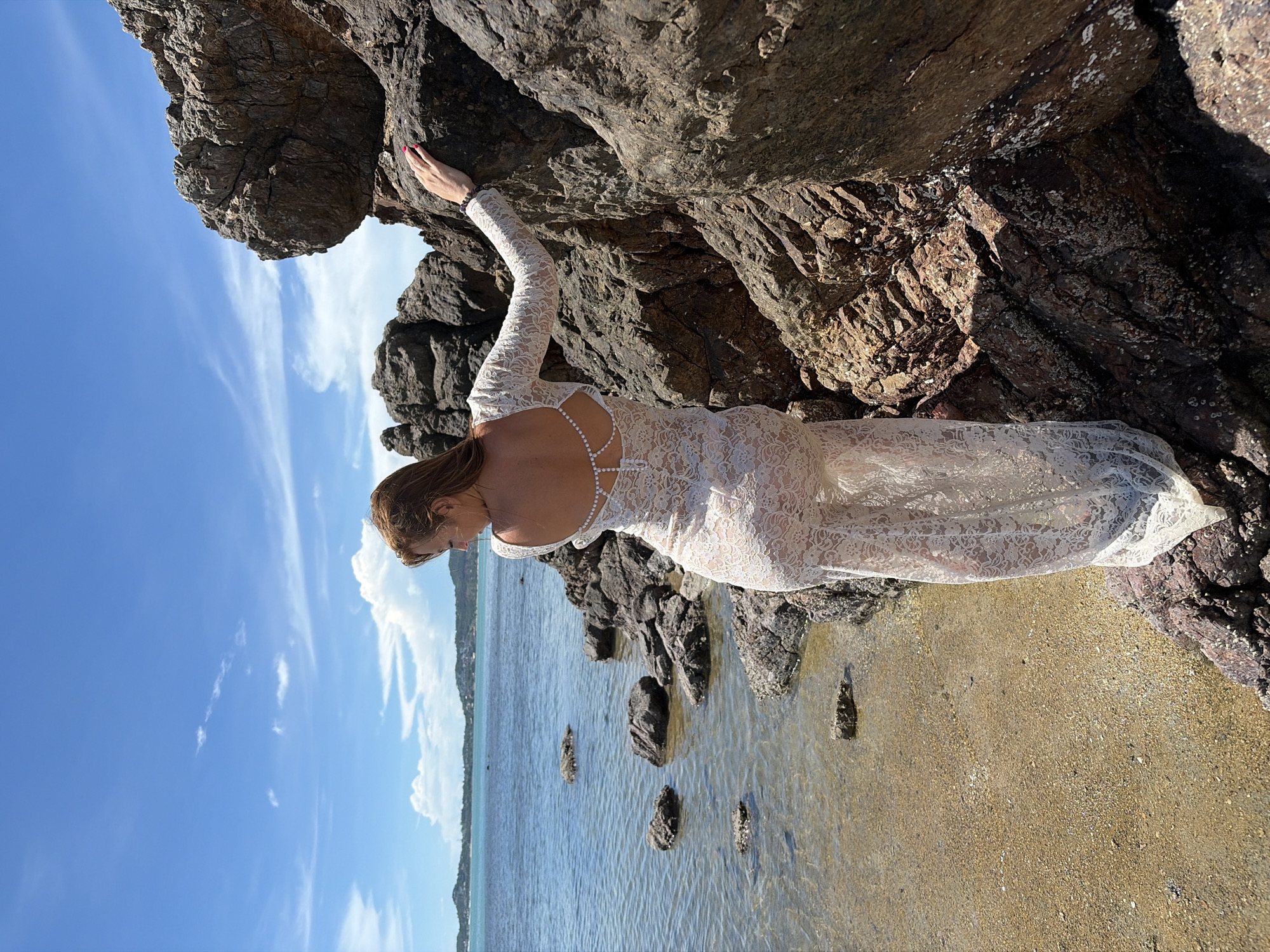 A woman in a white lace dress stands on rocky terrain near the water's edge, with her back to the camera. She is touching a large rock to her right. The water is calm and clear, with small waves lapping at the shore. In the distance, a coastline with buildings and vegetation is visible under a blue sky with scattered clouds. The scene appears to be a tropical beach.