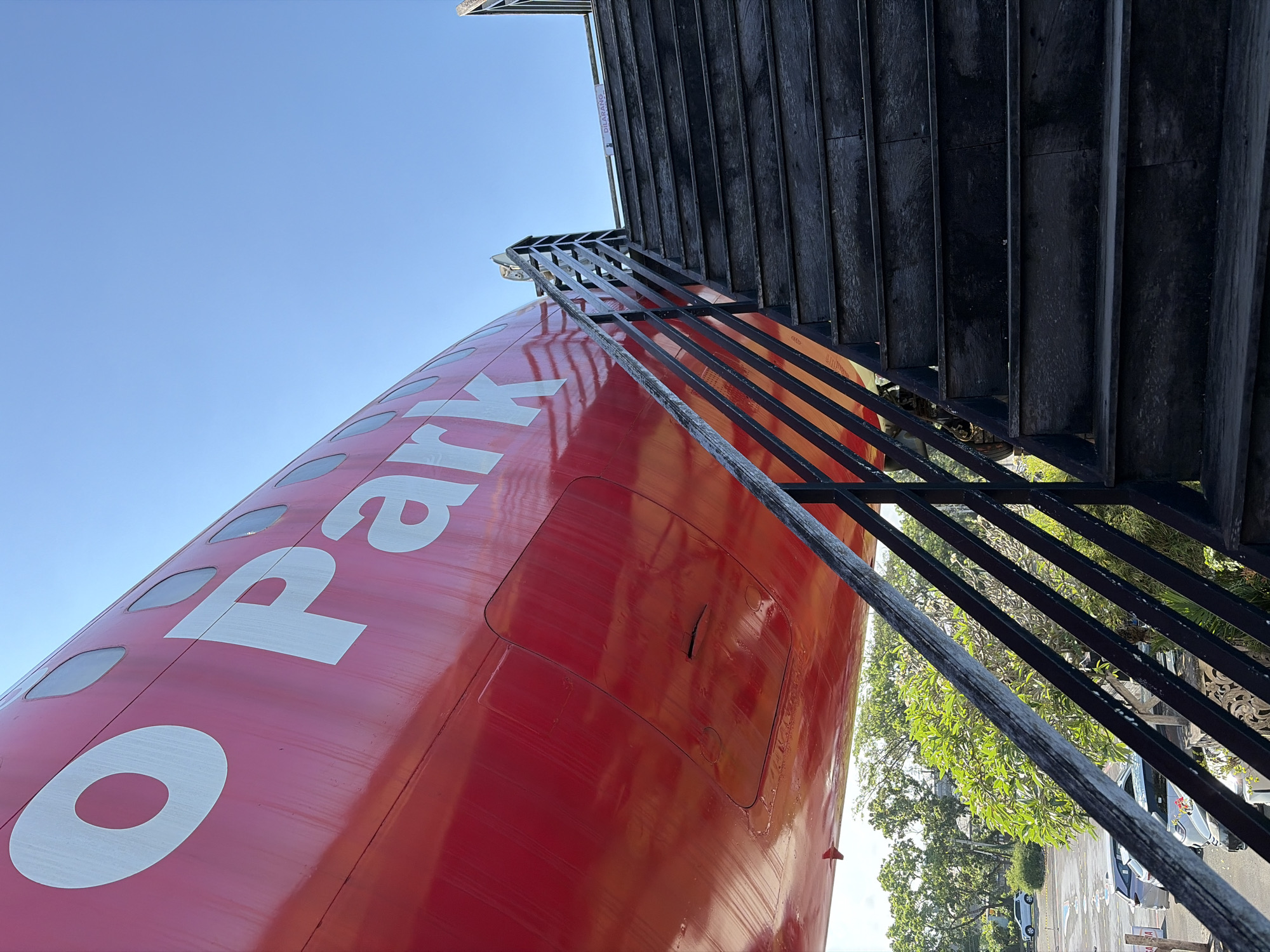 The image shows a red airplane fuselage, with the partial word "Park" visible in white lettering. A black wooden staircase with a metal railing leads upwards towards an opening in the plane. The stairs are worn and appear to be made of dark wood. Above the stairs, a sign with text in a foreign language is affixed to the railing. The sky is clear and blue. In the background, there are green trees and parked cars, suggesting an outdoor location. The context provided indicates this scene is in Blahbatuh, Indonesia.