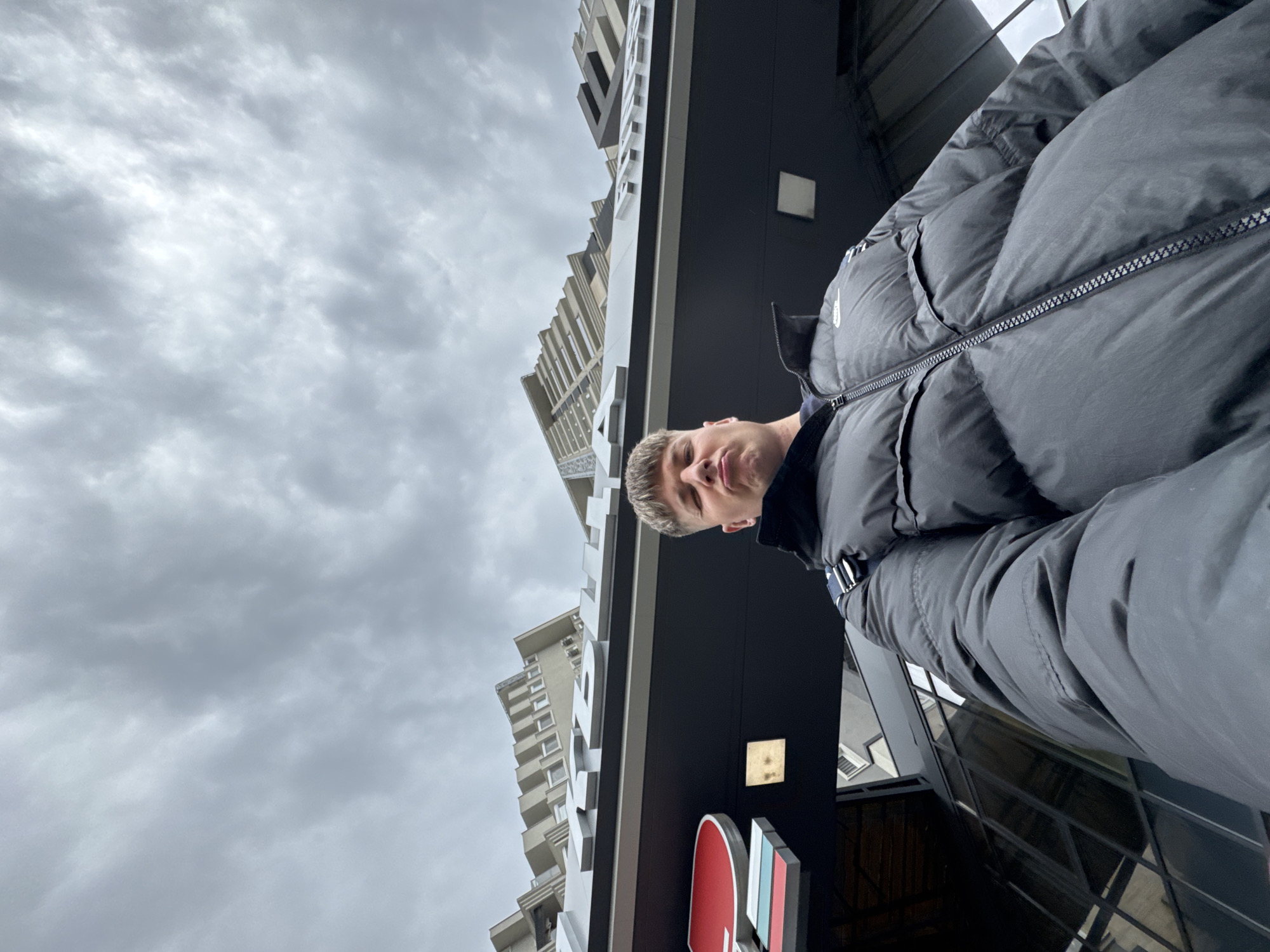 A low-angle medium shot shows a man in a black puffer jacket standing in front of a building in Plovdiv, Bulgaria. In the background, to the left of the man, is a tall building with white facades and windows. To the right and slightly behind the man, a portion of a building's black facade is visible, and part of a red, white, and blue sign. The sky is overcast.