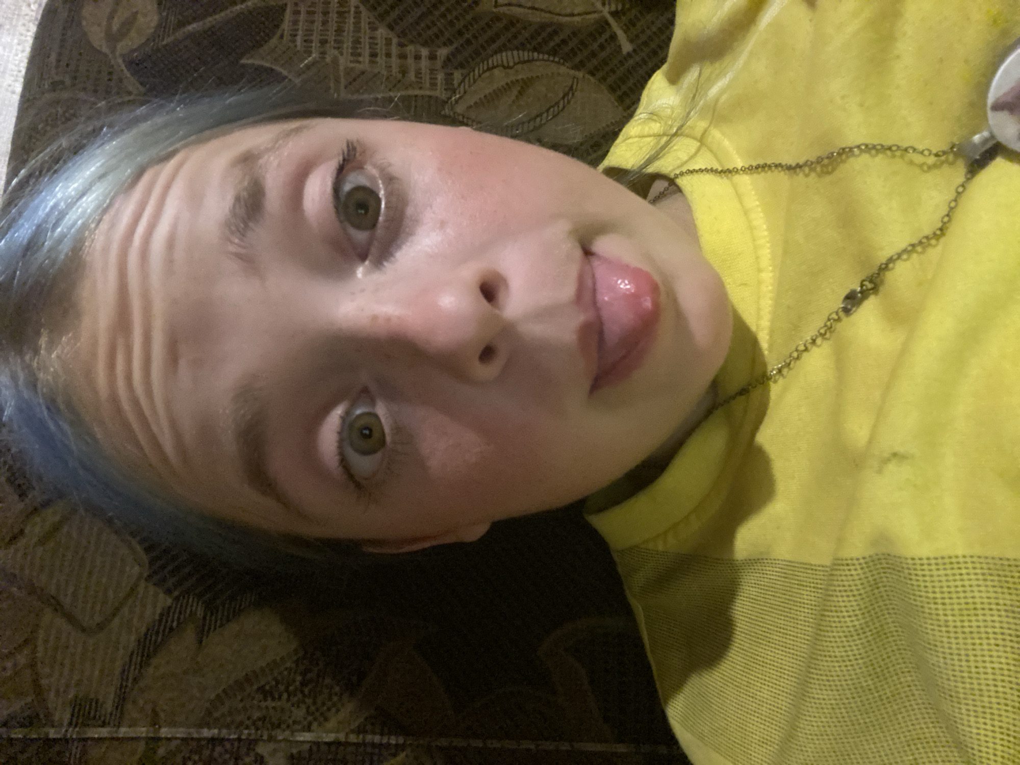 This is a close-up, Dutch angle, low-angle shot of a young person lying down. The person has light skin, green eyes, and blue-tinted hair. They are sticking their tongue out and have a playful expression. Their forehead is creased. They are wearing a bright yellow shirt with a striped pattern on the lower portion and a thin metal chain necklace with a round pendant. The background appears to be a brown couch with a textured pattern. The lighting suggests it's indoors, possibly in the evening. No visible text is present in the image. The location context is Nova Praha, Ukraine.