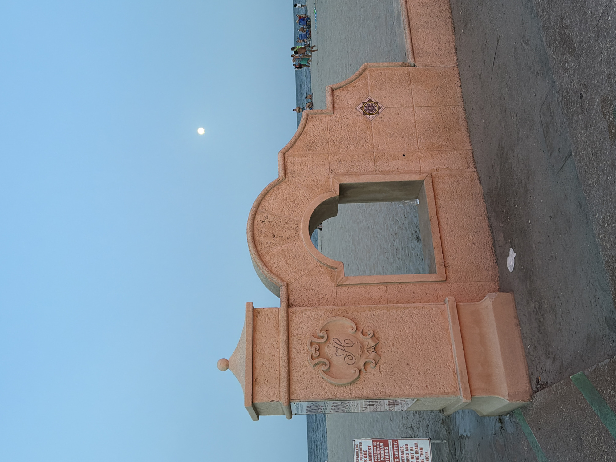 An outdoor scene depicts a decorative archway structure on a beach in Hollywood, United States of America. The archway, made of coral-colored material, features an arched opening looking out onto a sandy beach. A stylized "H" crest is visible on the left pillar of the archway, and a patterned tile is embedded on the right side. The sky above is a clear, pale blue with a full moon visible in the upper right quadrant of the image. The beach is populated with several people in beach attire, some sitting in chairs and others walking. The ocean is visible in the background. In the foreground, a concrete walkway with green painted lines is present. A sign with text in English and Spanish is attached to the left pillar of the archway, and some litter is scattered on the ground. The lighting suggests it is either dusk or dawn.