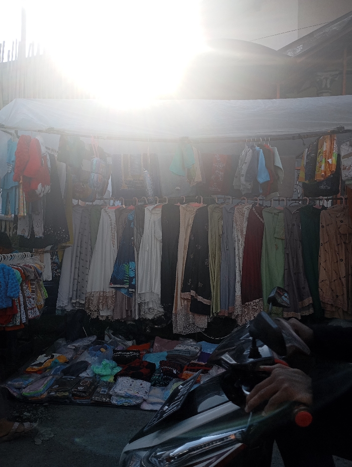 The image shows a street market stall in Kutawaringin, Indonesia (latitude: -6.974, longitude: 107.519). The sun is shining brightly, creating a hazy glare over the scene. A white awning shelters the stall, which is lined with rows of clothing items. A person's hand is visible, likely belonging to the vendor, resting on the front of a black scooter parked in front of the stall. There is an abundance of colorful clothing on display, and a variety of other merchandise is stacked and spread out on the ground in front of the stall. The image captures a typical scene of a bustling street market in a busy urban environment.