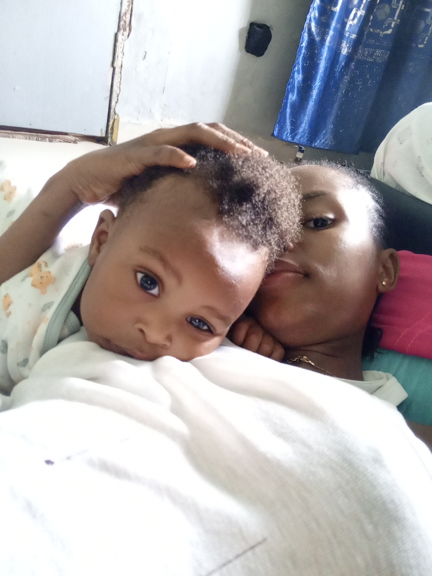The image shows a person and a baby lying down, possibly on a bed or a similar soft surface. The baby has curly hair and blue eyes. The baby wears a light-colored outfit with a patterned design. The other person has their hand on the baby's head. The background includes a blue curtain and a wall with what appears to be damaged paint or plaster. The location is Keffi, Nigeria.