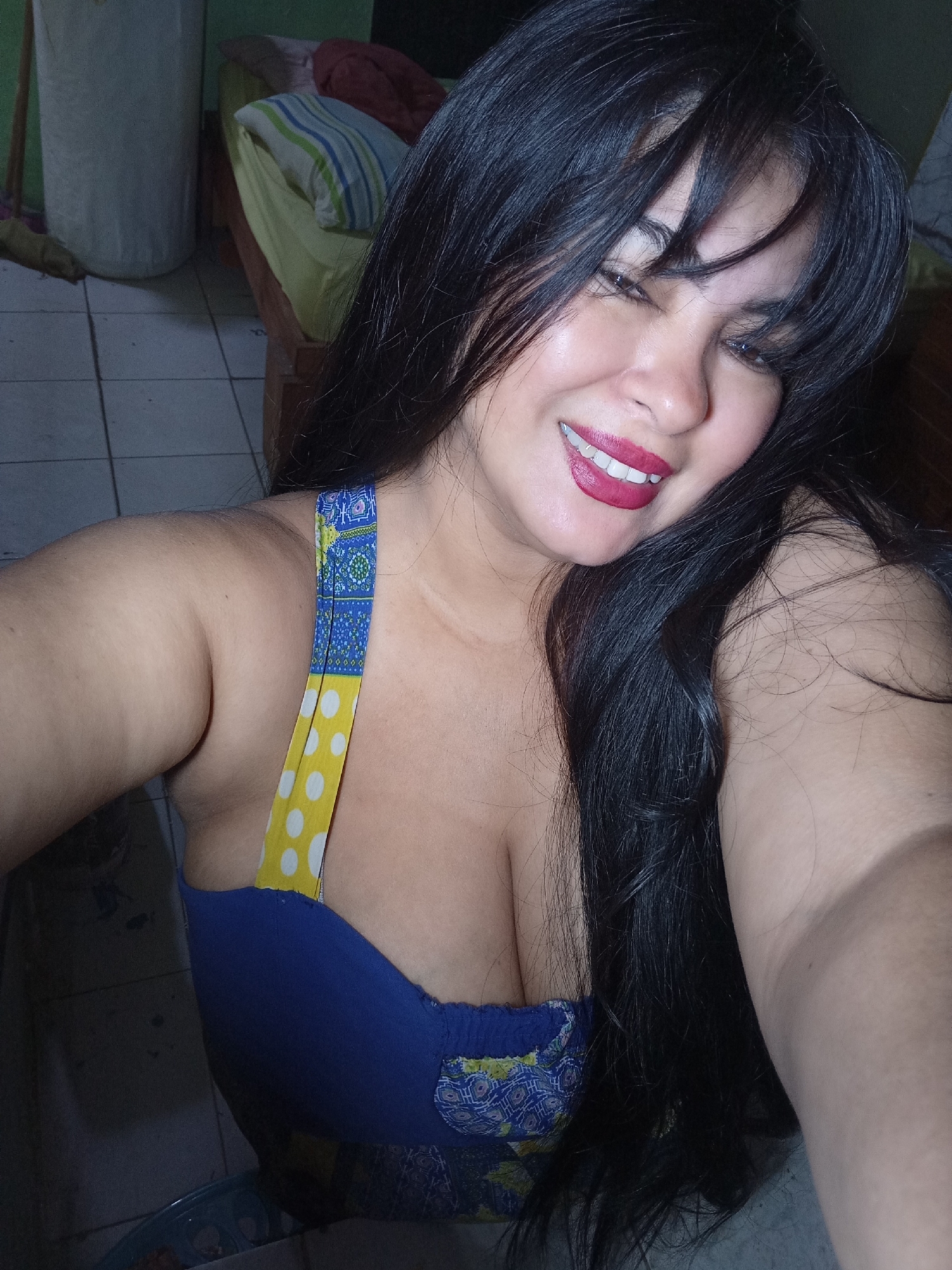 A woman is captured in a close-up selfie, smiling widely at the camera. She has long, dark hair, possibly with bangs, and is wearing red lipstick. Her skin appears warm-toned. She is wearing a blue top or dress with colorful straps; one strap features a yellow and white polka dot pattern, while the other has a blue and yellow intricate design. Her cleavage is visible.
The background suggests an indoor setting, likely a room with a tiled floor. To her right, a bed or cot with a light green sheet and a blue, green, and white striped pillow is partially visible, along with another reddish fabric. To her left, a large, light-colored cylindrical object, possibly a laundry hamper or a water container, stands against a wall. The lighting appears to be indoor, with her face being well-lit while the background is darker, suggesting a somewhat dim environment. The woman's expression conveys happiness and cheerfulness. No visible text is present in the image.