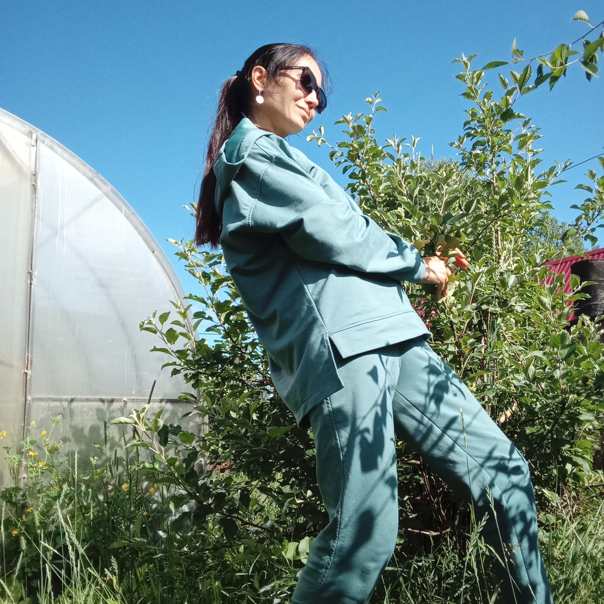A low-angle shot captures a woman outdoors on a sunny day, leaning against a lush green bush. She is wearing a teal tracksuit and sunglasses, with her dark hair pulled back in a ponytail. Her posture suggests she is reaching for something on the bush. In the background, a large, translucent greenhouse structure is visible to the left, and a red roof is partially visible to the right. Tall grass and other plants surround her. The bright blue sky indicates clear weather. The scene appears to be in a rural or garden setting, possibly a greenhouse area in Буренка, Russia.
