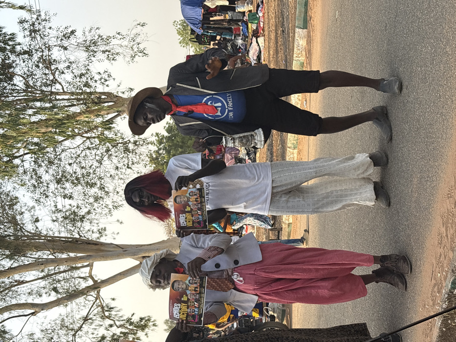 The image depicts three individuals in performative attire standing on an asphalt road in Lafia, Nigeria, alongside an open-air market under bright daylight.
From left to right:
1. A dark-skinned individual wears a white bonnet-like head covering, a light grey blazer over a checkered or plaid long-sleeved shirt with a red tie, voluminous pink trousers, and brown lace-up boots. White powder or paint is visible on the face. This person holds a glossy horizontal poster with both hands at waist level, displaying the title "ECHOES VICTORY" alongside multiple portrait photographs and the text "P.P.M" and "BALENCIAGA."
2. A second dark-skinned individual wears a black wig with red tips, dark eye makeup or paint, a plain white t-shirt, white loose-fitting trousers with perforated patterns, and grey slip-on shoes. This person also holds an identical "ECHOES VICTORY" poster horizontally with both hands at waist level, displaying the same content.
3. A third dark-skinned individual wears a straw hat, a dark blazer over a blue t-shirt featuring a white circular logo with "EF" or "LF" and "ECRA FAMILY," black shorts, and grey slip-on shoes. Red face paint or streaks are visible on the cheeks and forehead. A red bandana or scarf is tied around the neck. This person holds a silver-colored lantern in the left hand and gestures with the open right hand, palm forward, fingers spread.
The immediate foreground consists of asphalt road and a low green-painted concrete curb. Beyond the curb, reddish-brown soil is visible. The background features several tall, sparsely foliated trees against a clear, light sky. To the right and mid-ground, a bustling outdoor market is visible with various items, including textiles, spread on the ground. Several people are present, and portions of blue tarpaulin shelters are discernible.