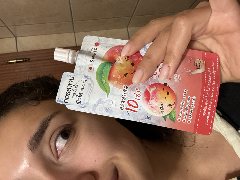 A female subject is partially visible from the mid-face upwards on the left side of the frame, smiling and looking directly into the camera. She has dark hair, dark eyebrows, and a brown eye. Her right hand, with manicured, natural-toned nails, holds a white, flexible pouch-packaging. The pouch features an illustration of peaches with cartoon faces, ice cubes, water droplets, and includes Thai script, the brand "SMooTO", a small Japanese flag icon, and the product name "Smooto Peachy Ice White Collagen Gel". The background consists of a light brown or beige tiled wall and a brown wooden door with a brass-colored hinge visible on the upper left. The specific location in Miljevci, Montenegro, cannot be determined from the visual evidence within the image.