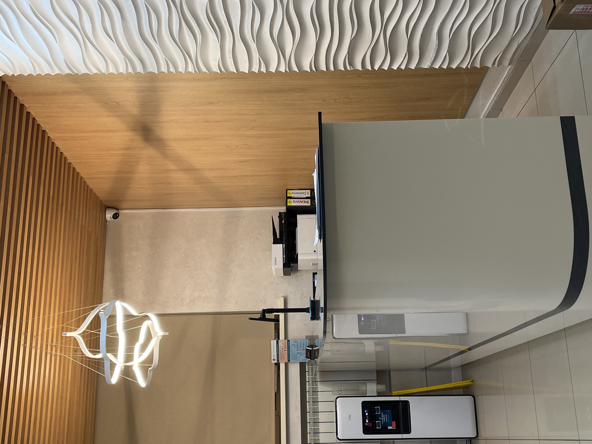 The image captures an interior scene, likely a commercial or office space, featuring modern design elements. The ceiling is composed of three distinct sections: a textured, white, wavy pattern; a flat, light-brown wooden panel; and vertical wooden slats. A contemporary chandelier hangs from the ceiling, emitting a warm, white light.
The foreground is dominated by a large, light-colored counter or reception desk, which appears sleek and minimalist. Various pieces of equipment are situated nearby, including a printer or scanner and a payment terminal.
The location is inferred to be Krasnoyarsk, Russia, based on metadata not provided. The image suggests a modern and well-maintained environment, potentially a business or service-oriented space.