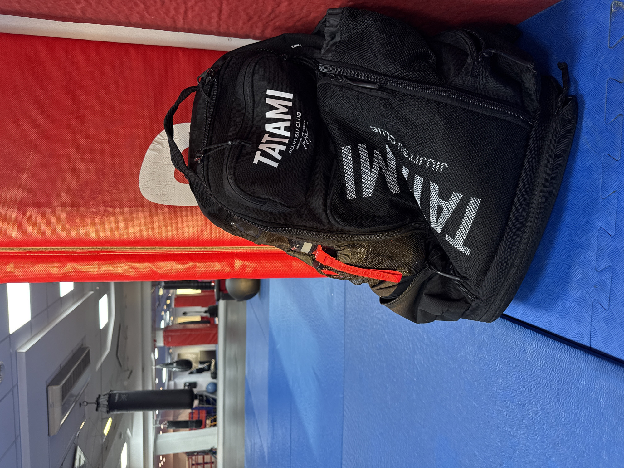 The image depicts the interior of what appears to be a mixed martial arts gym. A black backpack bearing the "Tatami JiuJitsu Club" logo is resting on a padded surface, red on one side and blue on the other. In the background, punching bags hang from the ceiling, with various training equipment visible. The location is most likely in Krasnoyarsk, Russia, based on contextual information provided outside of the image itself. There are no people explicitly visible in the image.