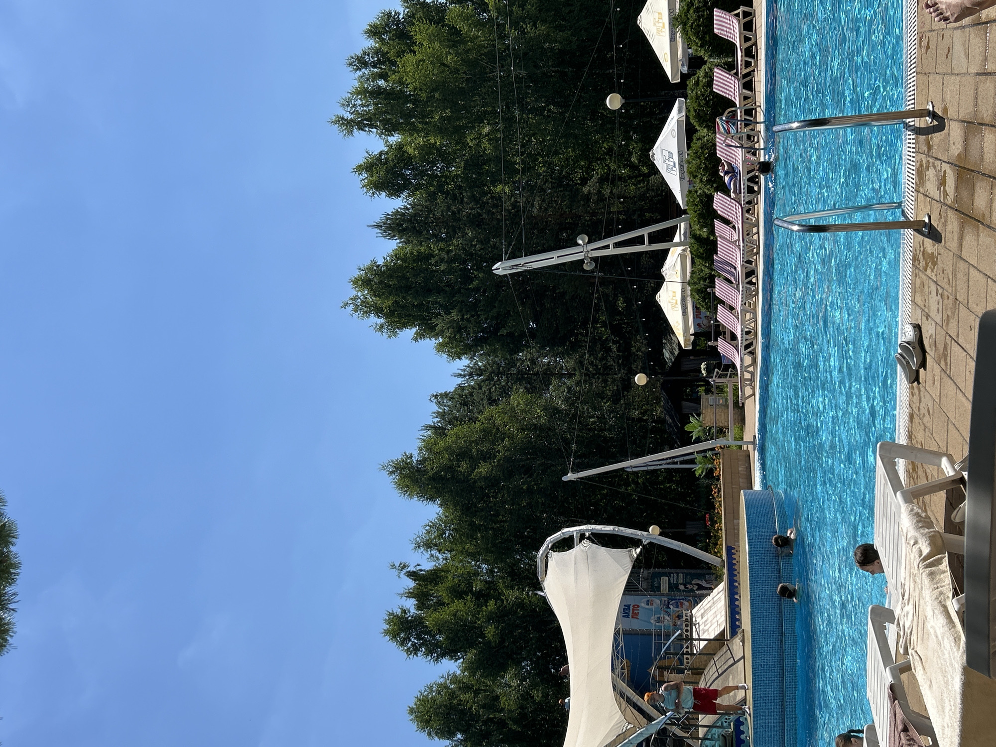 This image depicts an outdoor swimming pool area on a clear day, likely a leisure facility. In the foreground, several lounge chairs are visible, some with towels draped over them. The pool itself is filled with bright blue water, and several individuals are swimming or relaxing in it. To the left, a raised platform with a white canopy structure and some signage is present. A person in red shorts and a dark top stands on this platform. Behind the pool, a line of lounge chairs and umbrellas is arranged, with a dense backdrop of green trees. The scene suggests a recreational setting, possibly within the city of Togliatti, Russia, given the context provided.