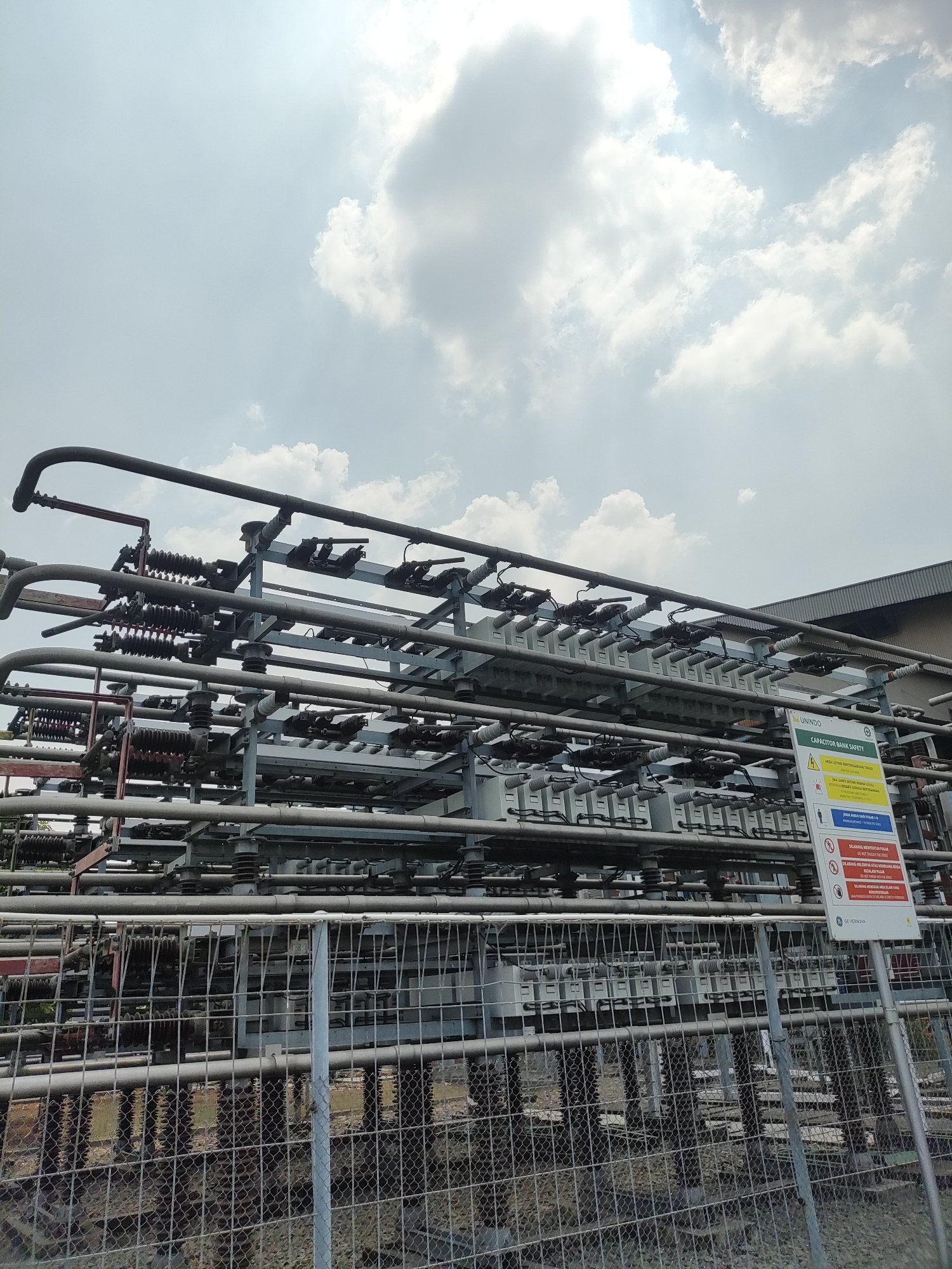 A medium shot, taken from a slightly low angle, captures an outdoor industrial scene on a partly cloudy day. The primary focus is a complex network of electrical equipment, featuring numerous metal pipes, insulators, and what appear to be capacitor banks, all mounted on a metal structure. A chain-link fence runs across the foreground, partially obscuring the base of the installation. To the right of the frame, a safety sign is prominently displayed, indicating "CAPACITOR BANK SAFETY" with various warnings in both English and what appears to be Indonesian. The background reveals a building with a sloped roof and a bright sky with scattered clouds, suggesting a sunlit environment. The overall impression is one of an active industrial site, likely a power substation or related facility, given the equipment and safety signage.