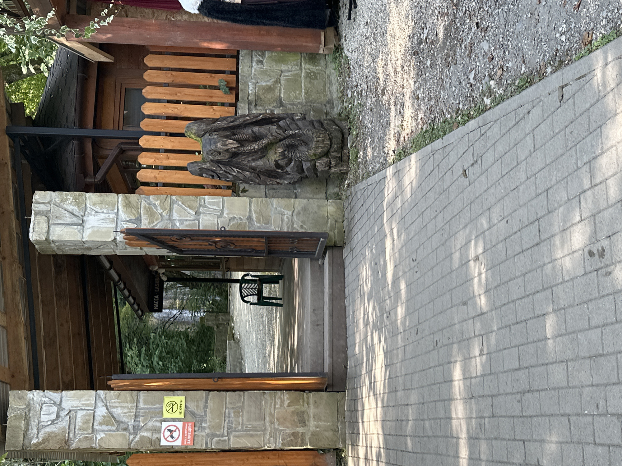 The image shows the entrance to a facility in Bol'shoi Kichmai, Russia. The entrance is framed by stone pillars and wooden gates. To the left, there are two signs: one with a prohibition symbol over a dog and text below, and another with a prohibition symbol over a cigarette and text reading "НЕ КУРЯТЬ". Inside the entrance, a green plastic chair is visible on a raised wooden platform. Further in, a sign reads "КАССА". To the right of the entrance, a wooden fence is present. A carved wooden sculpture of a winged creature entwined with a snake stands next to the fence. The ground in front of the entrance is paved with gray interlocking bricks. To the right of the paved area, there is a gravel path with some vegetation. The overall environment suggests an outdoor recreational area or a park entrance.