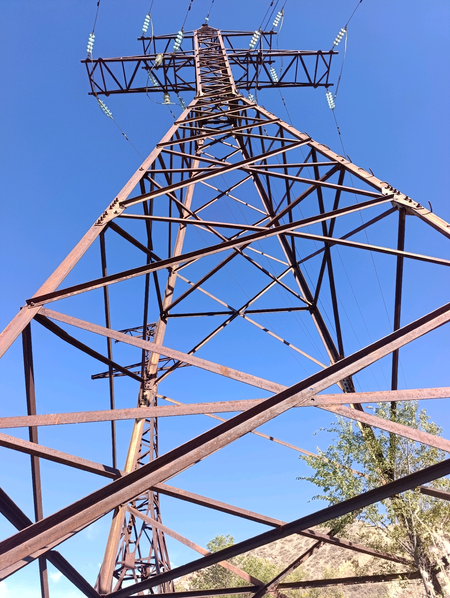 An upward-angle, full shot captures a massive, rusted metal transmission tower against a clear, vibrant blue sky. The tower is constructed from a lattice of crisscrossing steel beams, showing significant signs of corrosion and age. Several electrical wires, suspended by insulators, are visible extending from the top of the tower. In the lower right quadrant of the image, a portion of a leafy green tree and a dry, rocky hillside are visible, suggesting an outdoor, possibly rural or semi-rural setting. The overall scene is stark and industrial, contrasting with the natural elements present.