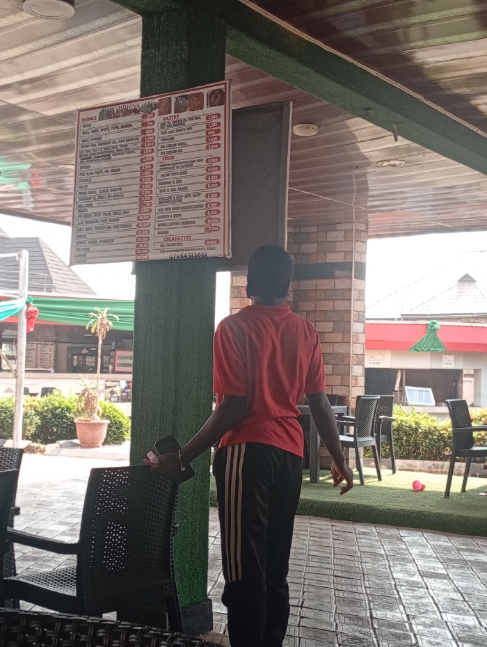 This image captures a bright, daytime scene at what appears to be an outdoor or semi-outdoor restaurant or cafe within the Akpaka Forest Reserve, Nigeria. The central figure is a person, seen from the back, wearing a red short-sleeved shirt and black track pants with three gold stripes down the sides, holding a phone in their left hand. They are standing in front of a prominent menu board, likely browsing the available items.
The menu board, hung from a ceiling beam, is divided into sections: "DRINKS," "PASTRY," "FOOD," and "CIGARETTES," with prices listed next to each item. Visible drink options include Water, Malt, Sprite, Pepsi, Mirinda, Fanta, and various alcoholic beverages like Amstel, Heineken, Gulder, and Johnnie Walker, with prices ranging from 500 for Water to 25,000 for Johnnie Walker. Pastry items include Cake, Doughnut, Meat Pie, and Egg Roll. The food section offers a variety of Nigerian dishes such as Fried Rice, Jollof Rice, White Rice, Salad with eggs, Moi Moi, Porridge, Yam & Egg Sauce, different "Swallow & Soup" combinations, Chicken & Chips, and Whole Catfish Barbecue (priced at 10,000). Cigarettes are listed at 2,000. A phone number, 07034513661, is displayed for complaints.
The setting includes dark, possibly rattan or plastic, chairs and tables arranged for dining. A green, textured material covers a pillar and frames the ceiling, which is made of brown corrugated or slatted material. The floor is tiled in a checkered pattern. In the background, other buildings, potted plants, and artificial grass are visible under a clear, sunny sky, indicating good weather.