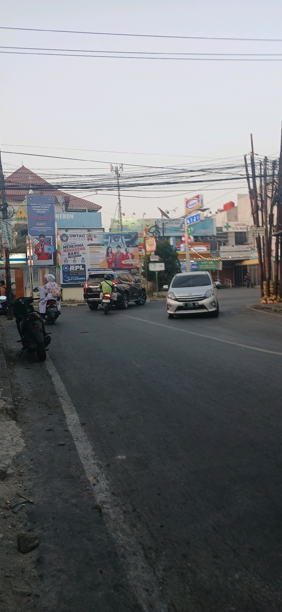 The image depicts a street scene in Cirebon, Indonesia, likely near a university campus given the prominent banners advertising UNTAG Cirebon for new student admissions. The foreground shows a road with a white line marking the lane. On the left side of the road, several motorcycles are parked along the sidewalk. A dark-colored SUV is stopped in the middle of the road, with a motorcyclist in a green shirt behind it. A silver car is approaching from the right side of the image. In the background, there are buildings with various advertisements and signs, including a prominent "Indomaret" convenience store sign. Overhead, numerous electrical wires are strung across the sky. The sky is overcast, suggesting it might be late afternoon or early morning.
