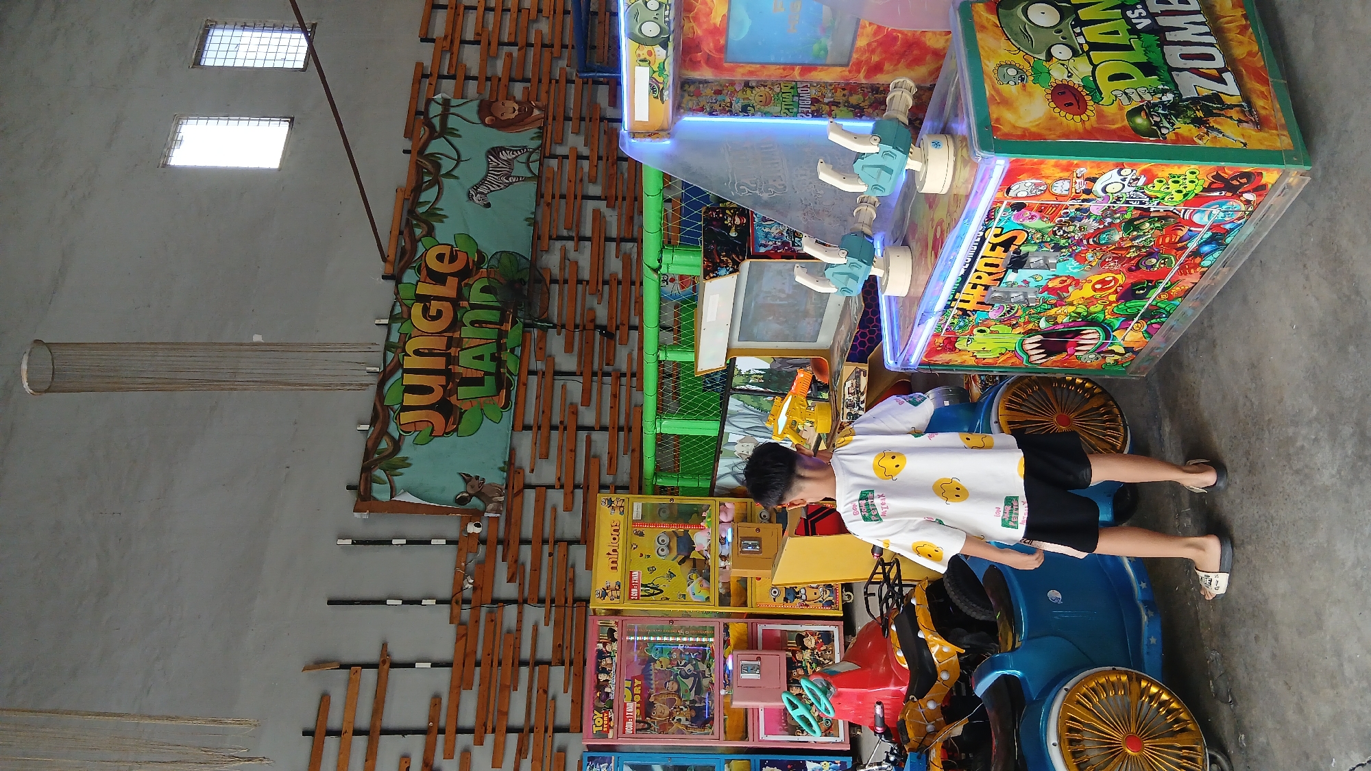 An indoor amusement arcade, likely in the city of Medan, Indonesia, features a young boy standing with his back to the camera, facing a row of arcade games. He is wearing a white t-shirt adorned with yellow smiley faces and black shorts, with light-colored sandals on his feet. The arcade environment is characterized by a concrete floor and a gray, textured wall. Above the games, a banner reads "JUNGLE LAND" with illustrations of jungle animals and foliage. The arcade machines include various colorful games, such as "Toy Story," "Minions," and a "Plants vs. Zombies" game, with a blue ride-on toy vehicle and a yellow toy motorcycle positioned in the foreground. The lighting suggests an ambient, possibly artificial, illumination.