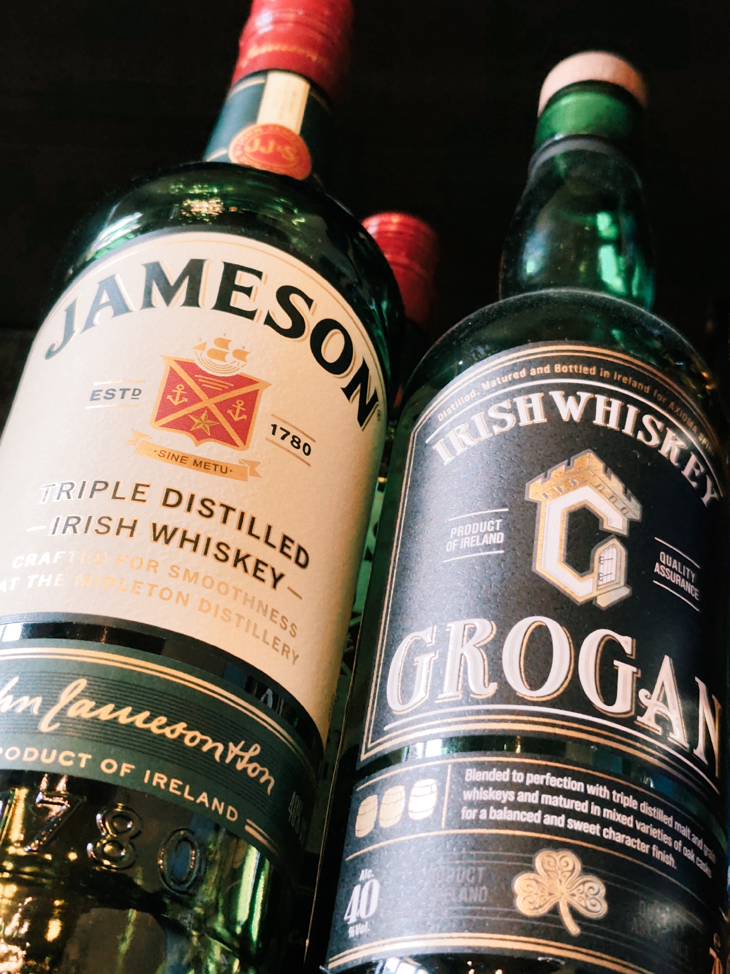 The image shows two bottles of Irish whiskey. The bottle on the left is Jameson, recognizable by its label. The bottle on the right is Grogan. Both bottles are made of green glass and have cream and black labels, respectively, with gold lettering. The image appears to be a close-up, likely taken indoors, possibly in a bar or liquor store. There is no indication of the city of Estosadok, Russia, in the image or its context.