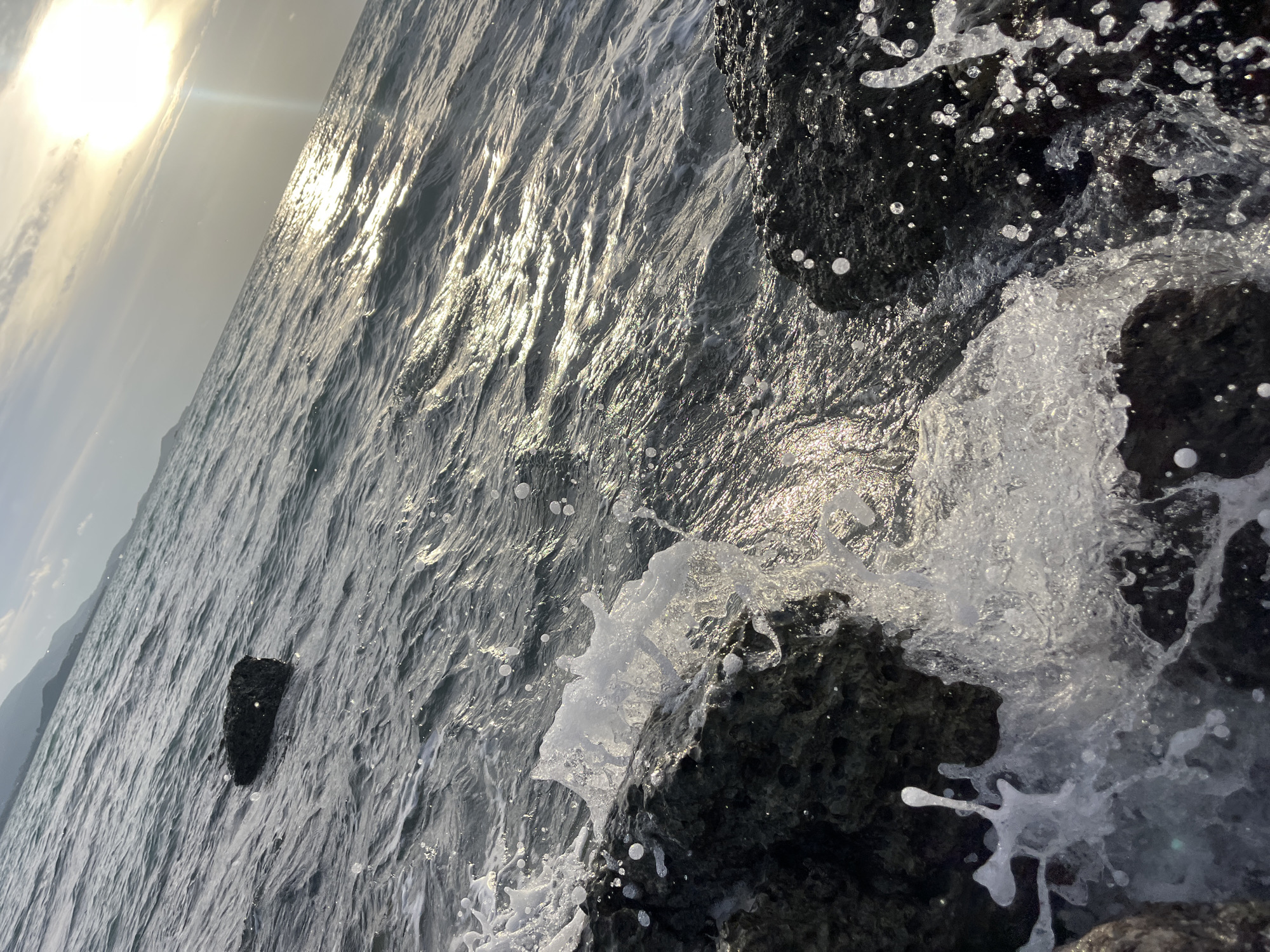 An oblique close-up shot captures waves crashing against dark, porous rocks on a coastline. The water is a deep, murky gray, with white foam and spray erupting as it meets the rocks. Sunlight glints off the water's surface, creating a shimmering effect. In the distance, a hazy outline of mountains and a cloudy sky suggest a coastal landscape, possibly in Ko Samui District, Thailand. A single dark, rectangular rock sits partially submerged in the choppy water in the mid-ground. The scene is devoid of any human presence.