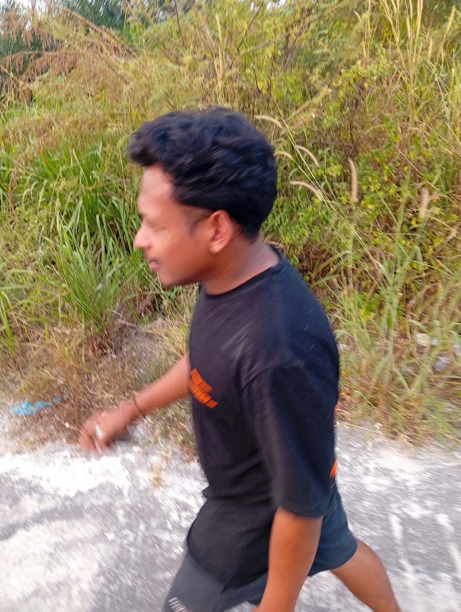 The image shows a medium shot of a young man in profile, walking along a paved path next to a grassy area. He is wearing a black t-shirt with an orange design and black shorts. The man has dark, wavy hair and is looking to his left. The background consists of dense, tall grass and some scattered foliage, with a bright, possibly overcast sky above. The lighting suggests it is daytime, with the sun likely positioned behind the camera. The paved path appears to be made of concrete, with some visible texture and discoloration. The overall scene appears to be an outdoor environment, possibly in a rural or semi-rural part of Kualu, Indonesia.