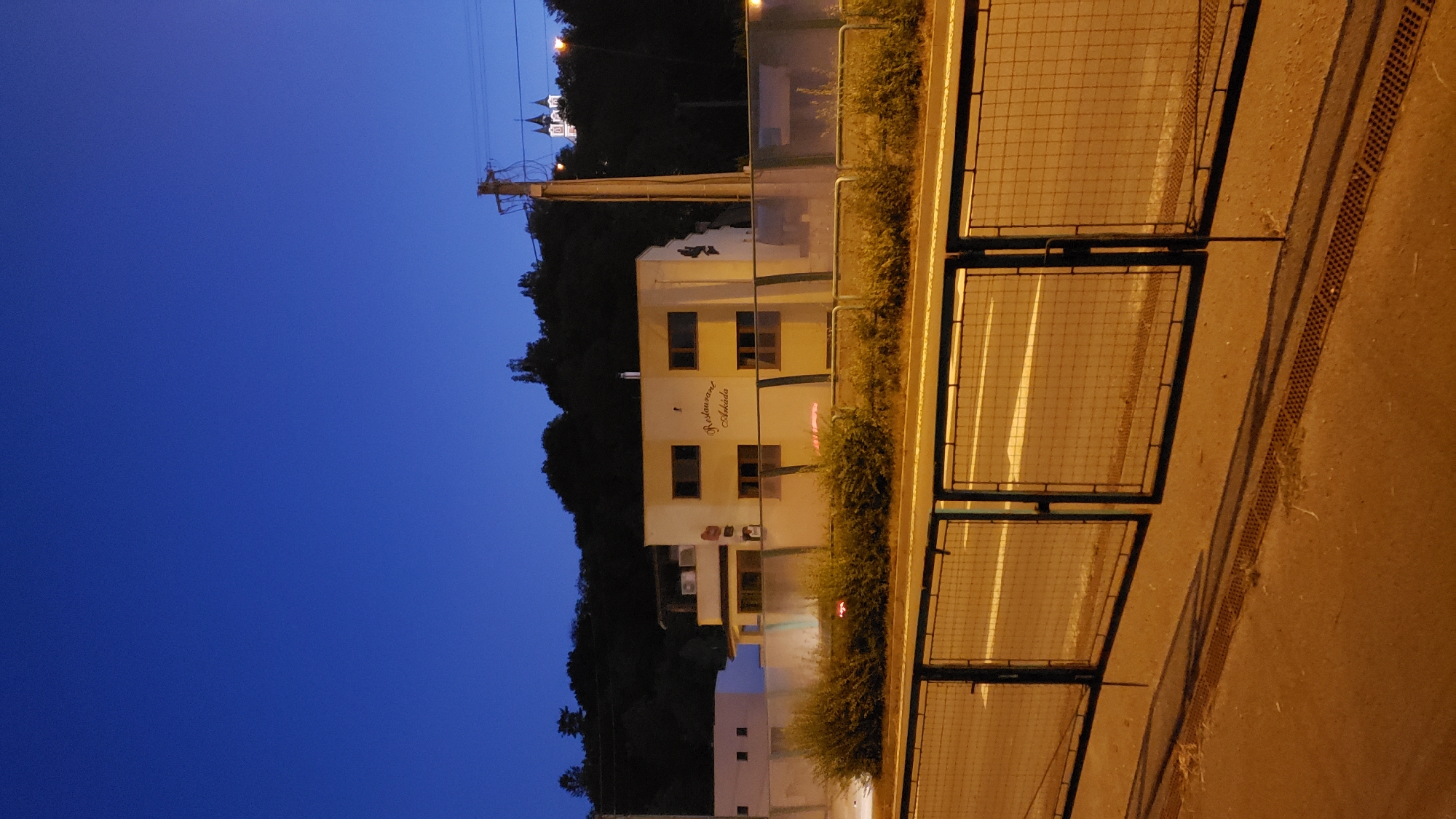 The image depicts a twilight scene in Prešov, Slovakia, featuring a modern building with the signage "Restaurant Arkada." The building is light-colored with multiple windows, and there are visible air conditioning units on an upper floor. In the background, a dark, tree-covered hill rises, and atop it, a distant church with multiple spires is illuminated. A utility pole with wires stands to the right of the building. In the foreground, a chain-link fence runs horizontally across the frame, separating the road from the building's property, which is bordered by dense green bushes. The road surface is asphalt, marked with white lines, and a drain runs along the edge. The sky is a deep, clear blue, indicating the transition from day to night. There are no people visible in the image.