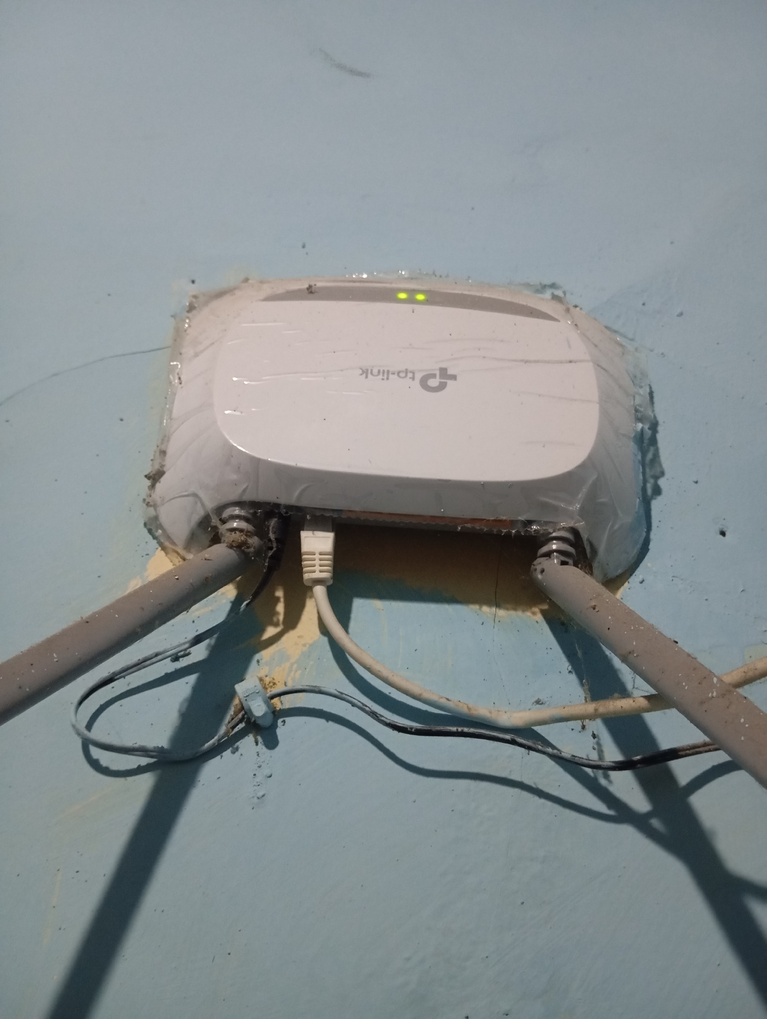 A TP-Link access point is mounted on a light blue wall. The device is wrapped in clear plastic sheeting. Two grey conduits enter the device from the bottom, angled upwards. A white Ethernet cable is plugged into the front of the device, and a black cable is also visible. There are two green indicator lights on the top of the access point. The wall has some yellow staining and a rough texture. The image appears to be taken in Sudikampiran, Indonesia, as indicated by the context.