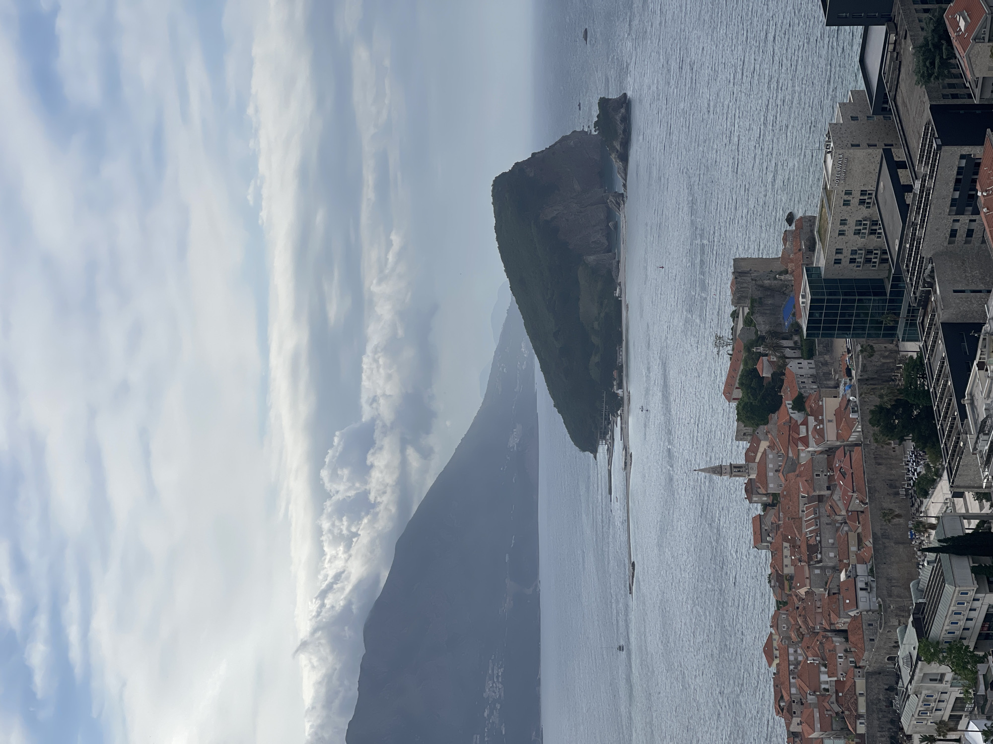 The image shows an aerial view of Budva, Montenegro. The Adriatic Sea stretches out, with a small island visible. The old town is characterized by red-tiled roofs and a distinctive church tower. Modern buildings are located on the outskirts of the old town. The sky is overcast with numerous clouds. In the background, a large, dark mountain emerges from the land and clouds.