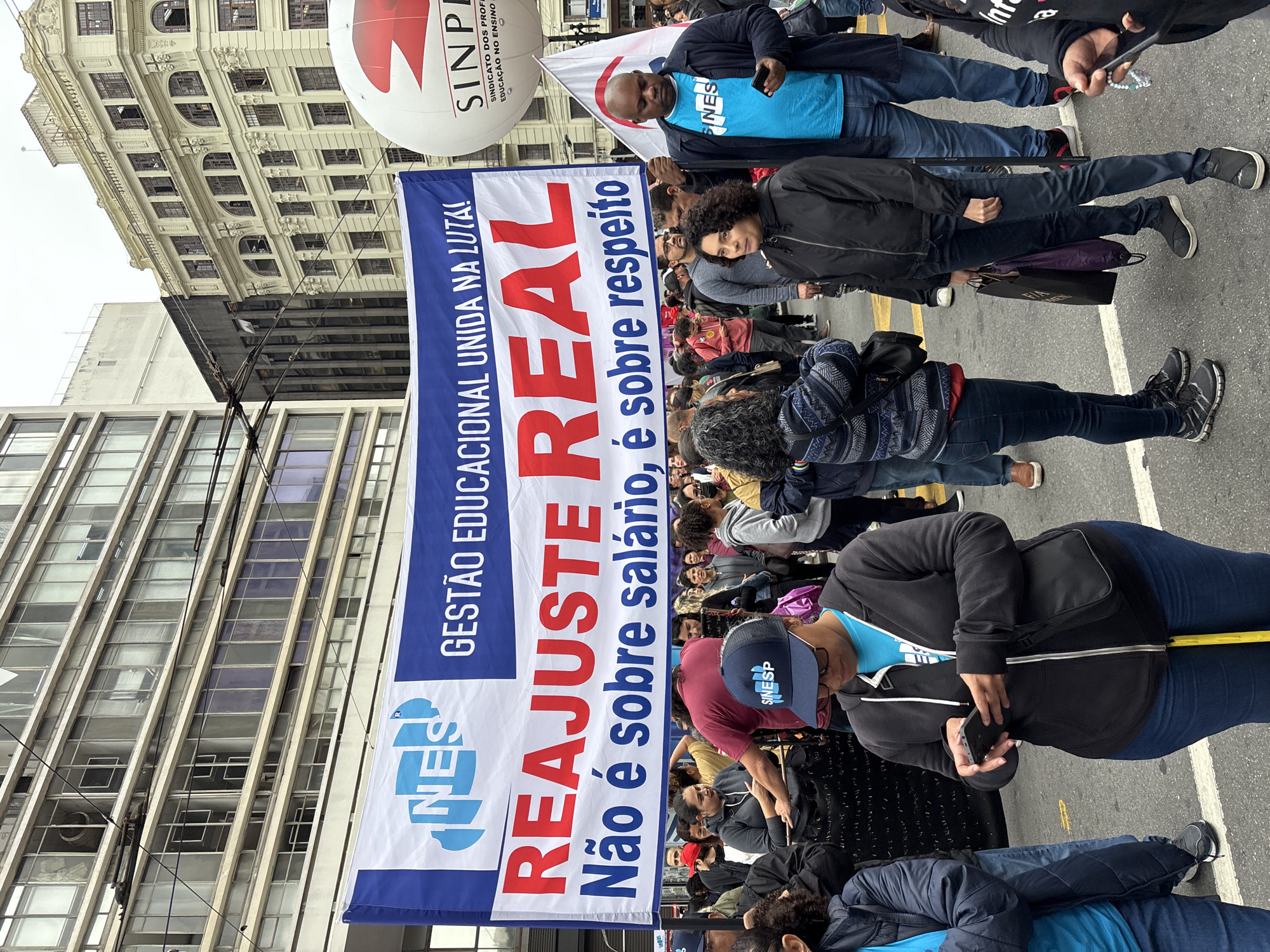 A street protest is taking place in what appears to be São Paulo, Brazil, where a group of people are marching, holding a banner with the inscription "GESTÃO EDUCACIONAL UNIDA NA LUTA!" (Educational Management United in the Struggle!), "REAJUSTE REAL" (Real Adjustment), and "Não é sobre salário, é sobre respeito" (It's not about salary, it's about respect). A SINPE balloon is visible. People are wearing casual clothes and holding phones, while some are looking at the banner and walking. Buildings form the background.
