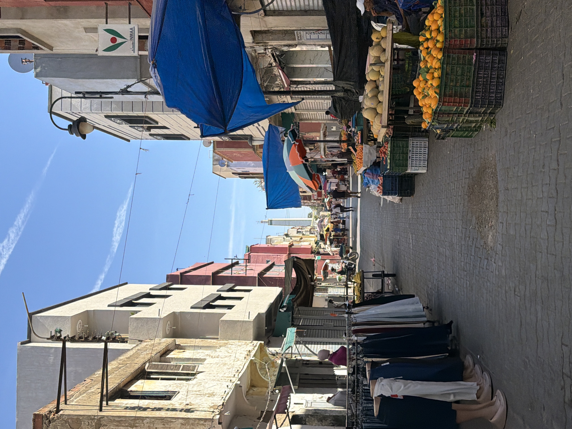 The image shows a street scene in Kenitra, Morocco, filled with market vendors and pedestrians. The narrow street is lined with buildings, some dilapidated, and others with modern architectural details. Blue awnings provide shade over the market stalls. Fruits, including melons and oranges, are displayed for sale. Clothing racks are visible on one side of the street. People are walking along the street, engaging in commerce. The sky is clear and blue, with contrails of airplanes visible. The street is paved with stone blocks. In the distance, a minaret can be seen rising above the buildings.