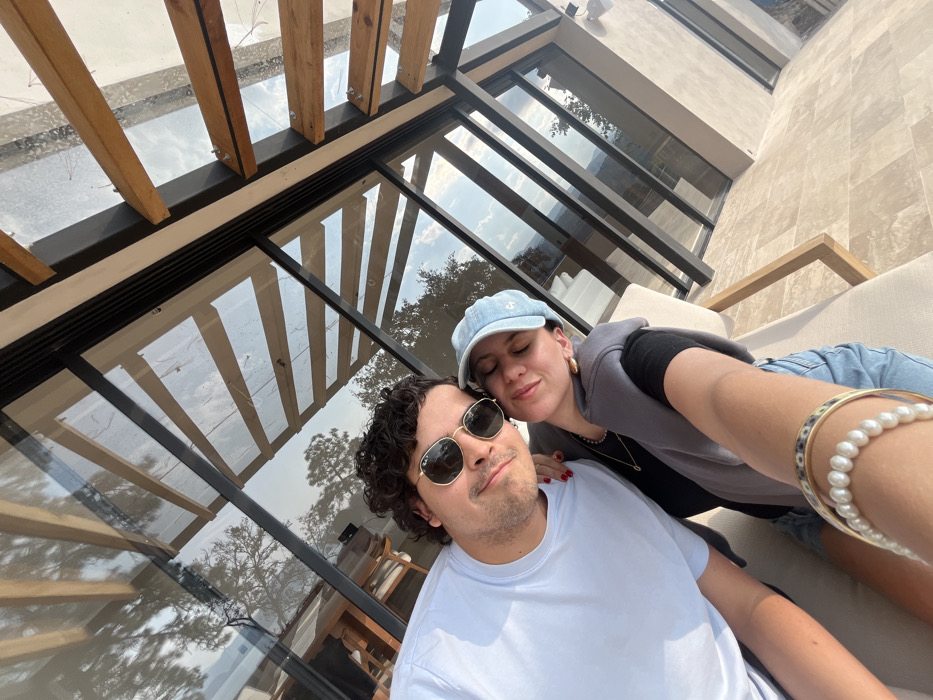 A couple poses for a selfie outdoors. The man wears sunglasses and a light blue shirt, while the woman dons a denim cap and gray hoodie. They are positioned against a background featuring a modern building with large glass windows and a wooden pergola above. Pearl and gold bracelets adorn the woman's wrist.