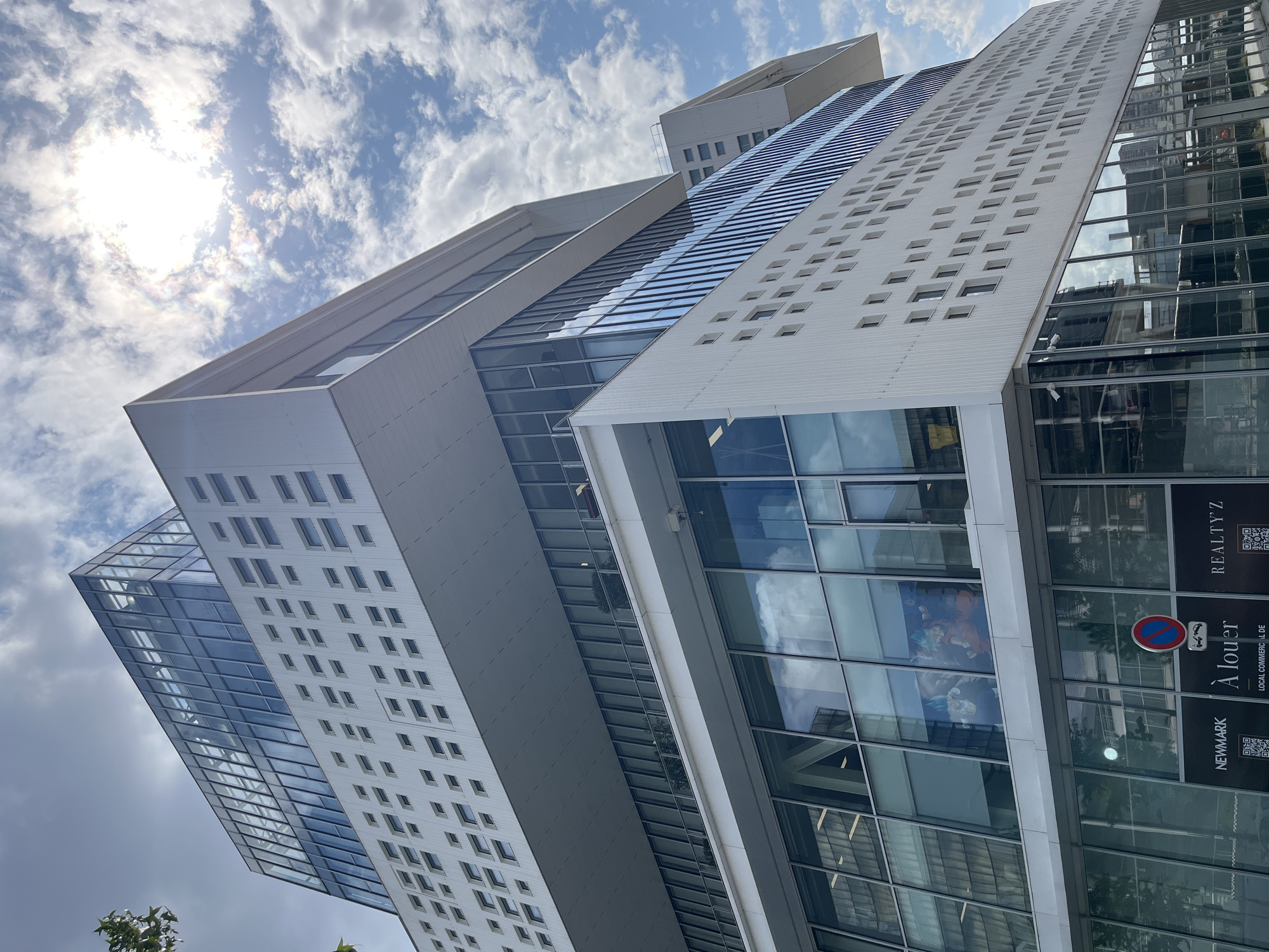 A low-angle, wide shot captures the facade of a modern, multi-story white building with geometric cutouts and large glass windows. The building's design features cantilevered sections and a glass-enclosed top floor. The sky is bright blue with scattered white clouds, and the sun is partially obscured by clouds, creating a halo effect. The lower portion of the building shows storefronts with signage including "NEWMARK," "À louer," and "REALTY'Z." Inside one of the glass-enclosed floors, a large mural or print of two anime characters is visible. A street sign with a red circle and a slash over a car symbol is present in front of the building. The surrounding environment appears to be urban, with reflections of other buildings visible in the glass facade.