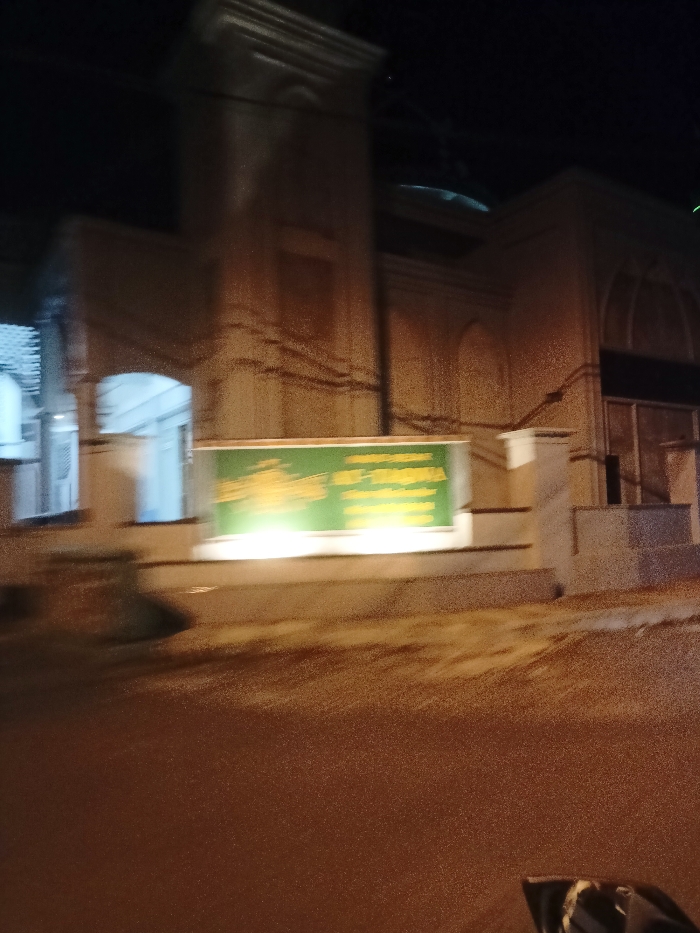 The image displays a building at night, illuminated by artificial lighting, against a dark sky. The architecture, featuring a prominent tower-like structure and visible dome, suggests it is a mosque. Arched windows or entrances are visible across the building's facade. In the foreground, a large, rectangular green sign with yellow lettering is brightly lit. The text on the sign is blurry due to low light and motion blur, but parts are discernible. The left section of the text appears to be in an ornate script, likely Arabic, possibly reading "Masjid" (Mosque). The right section contains Latin script, where "AGUNG" is visible, and the subsequent word closely resembles "MOJOSARI", indicating the sign likely reads "Masjid Agung Mojosari" (Grand Mosque of Mojosari), which aligns with the given location context of Mojosari, Indonesia. The foreground shows a paved ground surface, possibly a road, with reflections from the artificial lights. Steps are visible on the right side, leading up to an entrance. No people or specific activities are observed. The image captures a static, nocturnal view of a religious building.