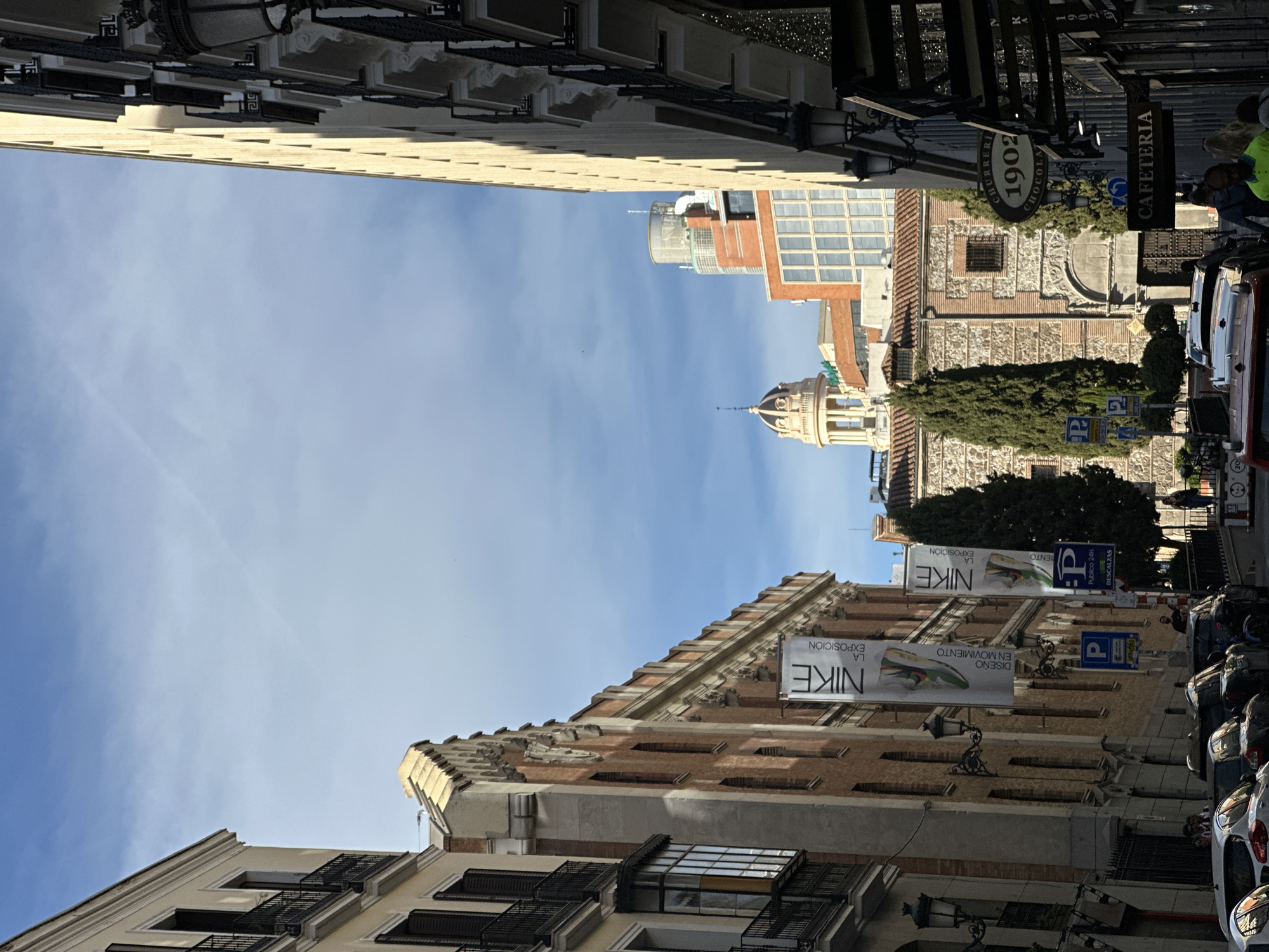 The image shows a view of a street in Madrid, Spain. Buildings line the street on both sides, with a clear blue sky visible above. One side of the street features a building with a 'NIKE LA EXPOSICIÓN' banner. Towards the end of the street, on the right side, there are signs for "Cafeteria", a "Churreria" sign that reads '1902', and a building with distinctive architecture, appearing to be a historical site. Cars are parked along the right side of the street. Several people are near the end of the street, towards the right.