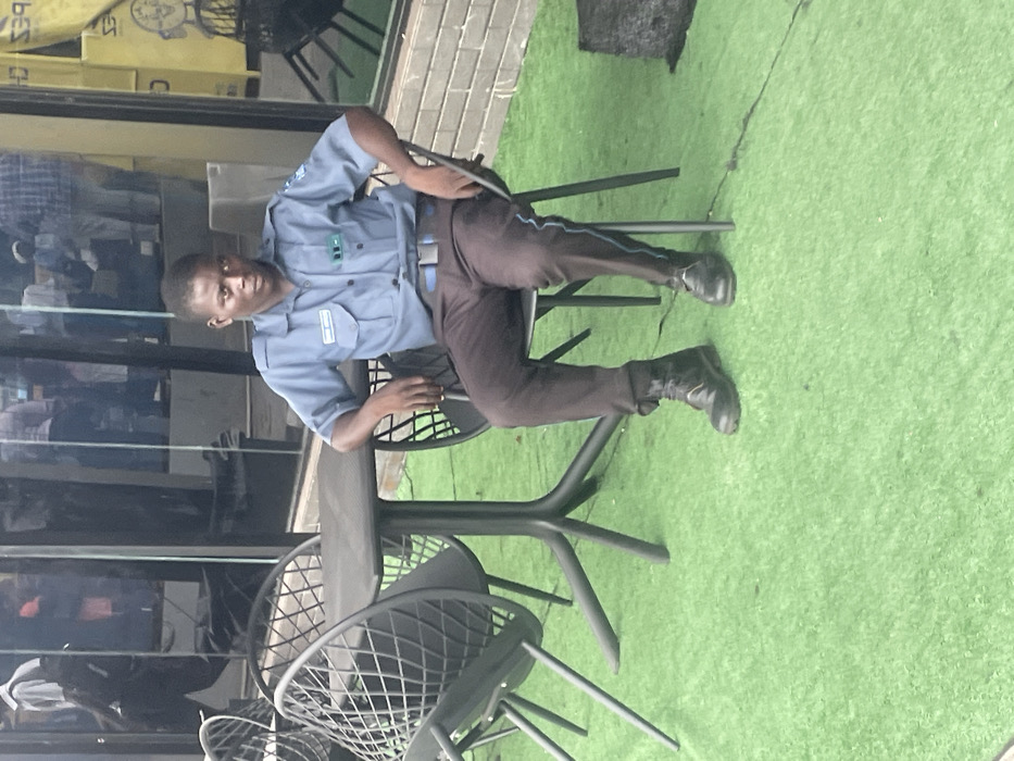 The image displays an outdoor seating area in Ikeja, Nigeria. A male individual is seated, facing forward, on a dark grey metal chair featuring an open geometric pattern on its back and seat. He is dressed in a light blue short-sleeved collared shirt with a "SECURE NIGERIA" patch on the left chest and a blue belt. He wears dark brown trousers, a black combat boot on his left foot, and a black dress shoe on his right foot. A thin blue stripe runs vertically down the outside of his right trouser leg. His arms rest on the chair's sides, and he maintains direct eye contact with the camera. To the left of the seated individual is a dark grey rectangular table with a central pedestal base branching into four legs. Another similar dark grey metal chair is partially visible to the left of the table. The ground surface consists of green artificial turf. In the background, a building with large glass windows reflects multiple indistinct figures and urban elements. A yellow sign, partially showing "CRAPES CH", is discernible through or behind the glass on the right side.