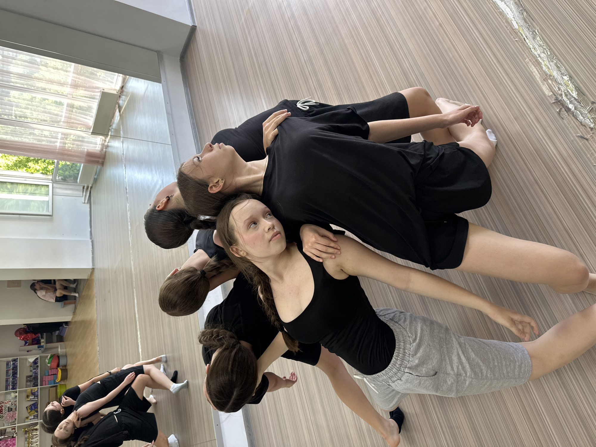 This media file captures a group of young women, likely dancers, in a studio setting. They are arranged in a flowing, interconnected pose, lying and leaning on each other. The subjects are all wearing black tops, with some wearing black shorts and others grey sweatshorts. Their hair is mostly tied back in braids or ponytails, suggesting a dance or practice session.
The scene is a dance studio, identifiable by the polished wooden floor and large mirrors on one wall, which reflect the ceiling and part of the room. The lighting suggests it is daytime, with natural light streaming in from an unseen window, illuminating the space. The overall mood appears focused and possibly relaxed, given their intertwined positions. There is no visible text in the image. The specific location is Votkinsk, Russia.