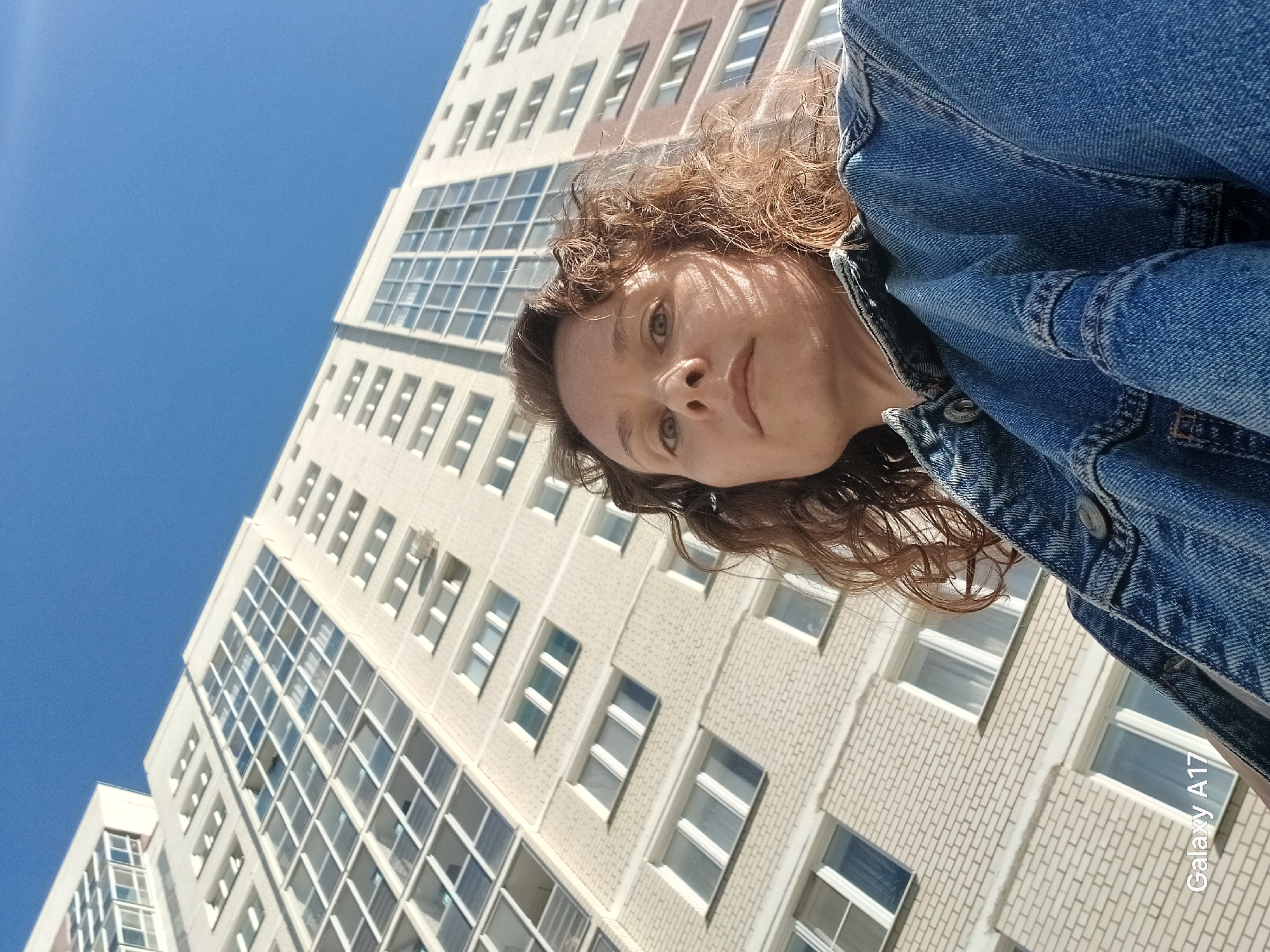 A low-angle photograph captures a woman with curly reddish-brown hair, wearing a blue denim jacket. The background features a tall, light-colored building with multiple windows, framed against a clear blue sky. The image includes the text "Galaxy A17" in the bottom right corner. Based on the architectural style of the building and the presence of Russian text in the file metadata, the location is likely Yekaterinburg, Russia.
