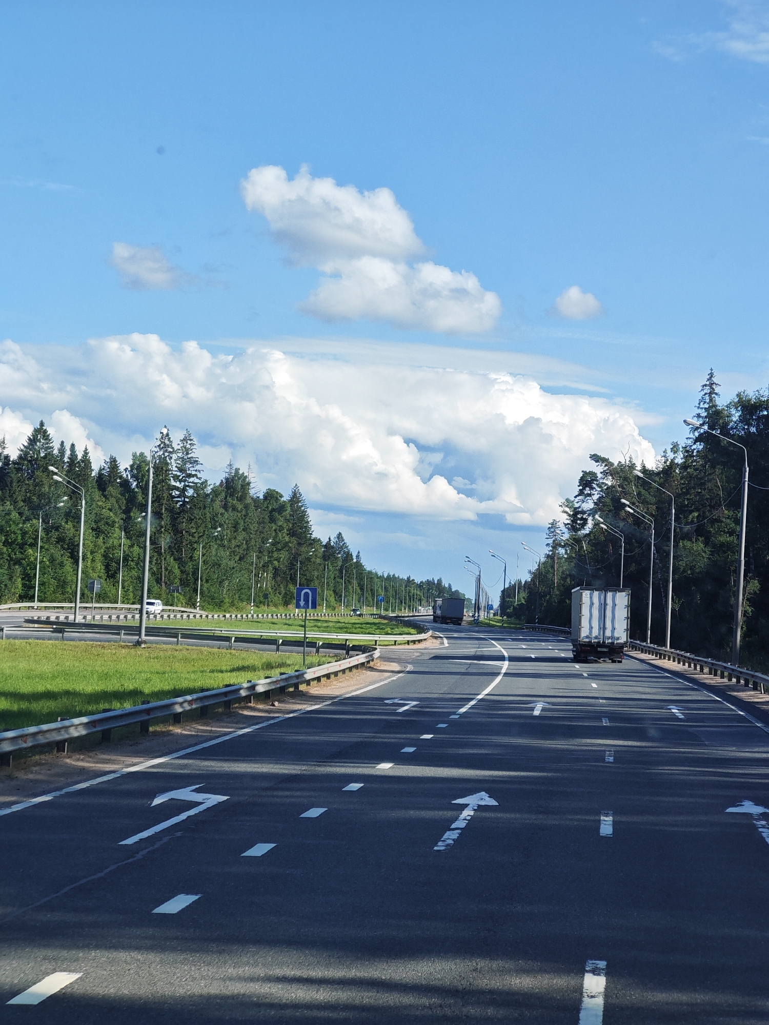 The image depicts a highway in Kuzhlevo, Russia, under a partially cloudy blue sky. The highway has multiple lanes with white lane markings, including directional arrows indicating traffic flow. Guardrails line the sides of the road, separating the lanes from grassy areas and a dense forest. Several streetlights are visible along the highway. In the distance, a white truck with a cargo container is traveling on the highway, and further ahead, another truck is visible. The scene is set during daylight hours.