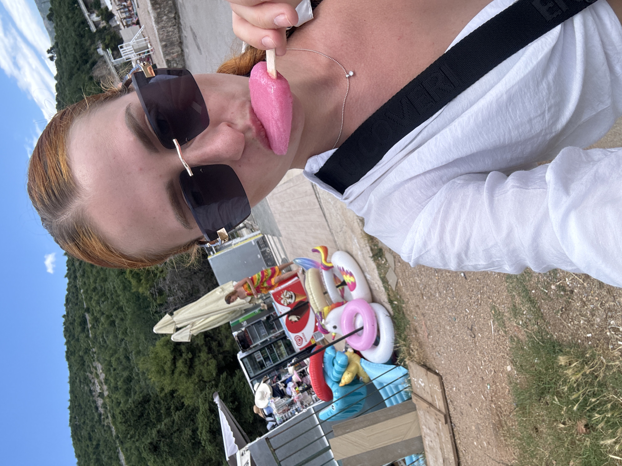 A young woman with auburn hair, wearing sunglasses and a white top, is taking a selfie while eating a pink popsicle. She is standing outdoors, with a lush green hillside and a blue sky with some clouds in the background. To her left, there are beach inflatables, umbrellas, and a cooler. In the background, there are buildings and a beach. The scene is likely in Budva, Montenegro, given the landscape and beach resort setting.