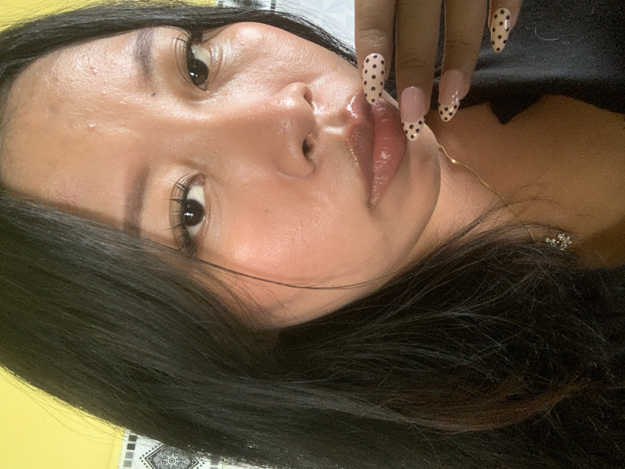 A close-up, waist-up shot shows a young woman with long, black hair. She is looking down and to her right, her mouth slightly open. Her lips are glossy and her eyes are dark. She has long, false eyelashes and her eyebrows are well-defined. Her skin appears to be slightly oily. She is wearing a gold chain necklace with a small, sparkling pendant. Her right hand is raised to her face, with her fingernails painted with a light pink base and black polka dots. The background is blurred, but appears to be indoors with a yellow wall on the left and a patterned surface on the right. The specific location in the city of Jajag, Indonesia cannot be determined from the visual information provided.