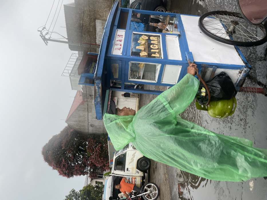 A man wearing a green raincoat is standing in the rain, pushing a food cart. The cart is blue and white, with a sign that reads "BASO LIGAR" in black letters. There are glass bottles and a can on the counter, with a few items inside. The cart has one wheel that can be seen, as the other is out of the frame. The man's hands are on the handle of the cart, and he is looking to the right side of the frame. The background of the image shows a grey sky, an obscured green tree and buildings in the distance. There are multiple puddles on the wet pavement. The man is wearing a black baseball cap, and his face is partly visible. This scene is in Ngamprah, Indonesia, at the coordinates -6.866 latitude and 107.516 longitude.