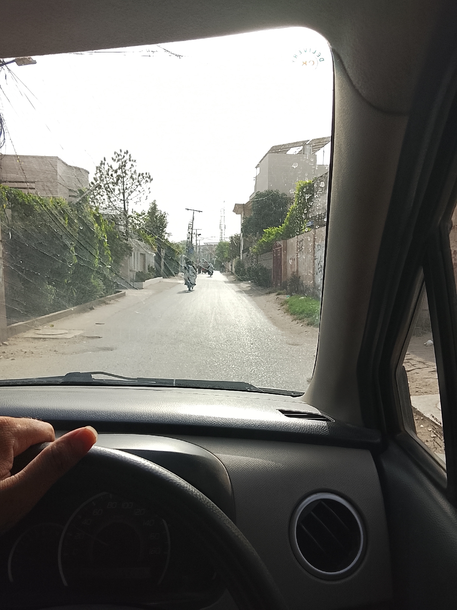 The image shows a view from inside a car driving down a residential street. The street is lined with buildings and greenery, including trees and vegetation growing over walls. Several motorbikes are visible in the distance. The driver's hand is visible on the steering wheel. The dashboard and air vent of the car are also prominent in the foreground. The sticker on the upper right corner of the windshield says, "DELIVERED." The location is identifiable as Gulgasht Colony, Pakistan, due to the typical residential street layout.