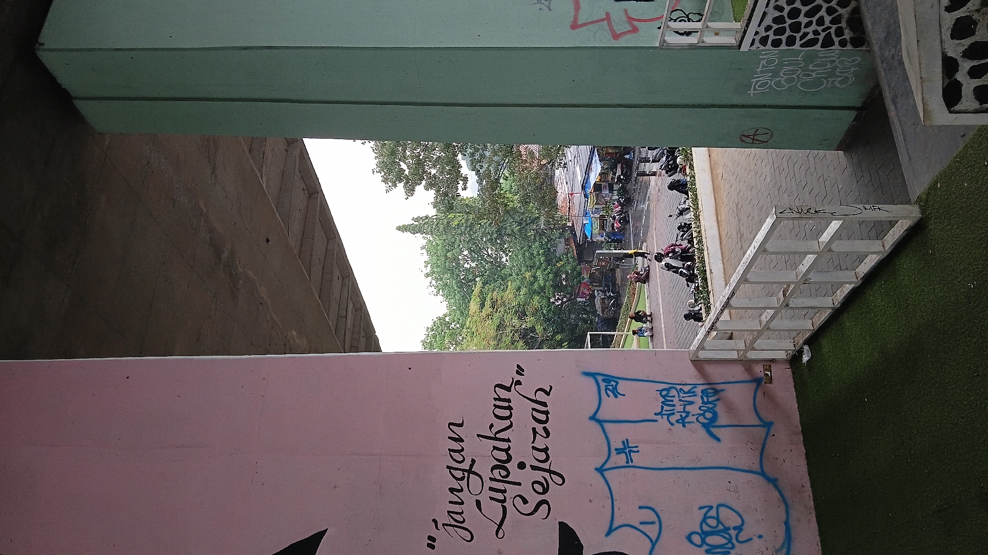 The image captures an outdoor urban scene, likely in Bandung City, Indonesia, under an elevated structure. On the left, a large pink pillar is covered with graffiti. In the center, a green pillar stands, also with graffiti on its surface. Between these pillars, a view opens up to an area with trees, market stalls, and several motorcycles. People are present, some sitting or standing near the stalls, while others are on or around motorcycles. The ground is paved with interlocking bricks, and a white railing separates the paved area from a section covered in artificial green turf. The sky is overcast, suggesting it might be a cloudy day or early morning.