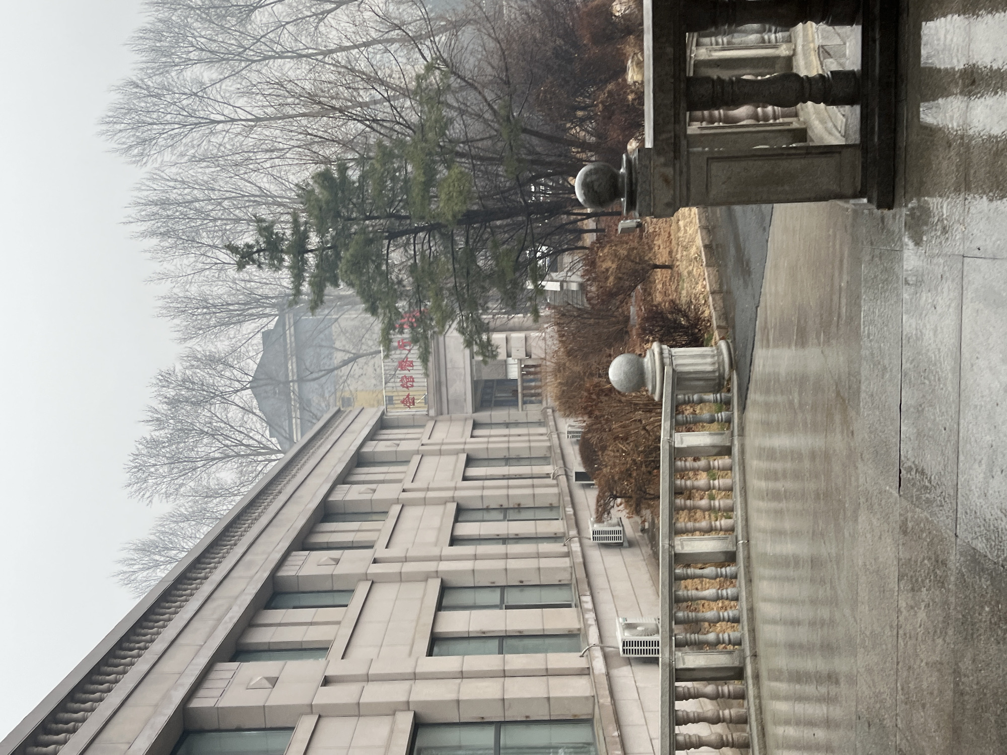 A photograph depicts a multi-story building in Changchun City, China, observed on a foggy day. The building is constructed with light-colored, possibly stone, blocks with several uniform window rows and horizontal details. Bare trees and evergreen foliage partially obscure the building's upper floors. There are also some words on the building in red lettering. In the foreground, there is a balustrade. The ground is wet, indicating recent rain.
