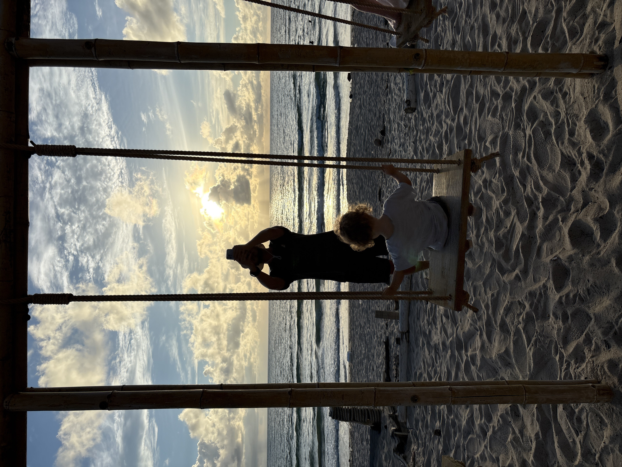 This media file shows a father and son on a beach in Choeng Thale, Thailand. The father is silhouetted against the setting sun, holding up his phone to take a picture. His son is sitting on a wooden swing, looking out at the ocean. The sand is rippled, suggesting the tide has recently receded. The sky is filled with dramatic clouds, illuminated by the low sun. The overall mood is peaceful and serene, with a hint of joy in the father's action. The image is framed by bamboo structures, likely part of a beach swing set or cabana. The time of day is clearly late afternoon or early evening, as indicated by the low angle of the sun and the golden light. The weather appears to be clear with scattered clouds.
