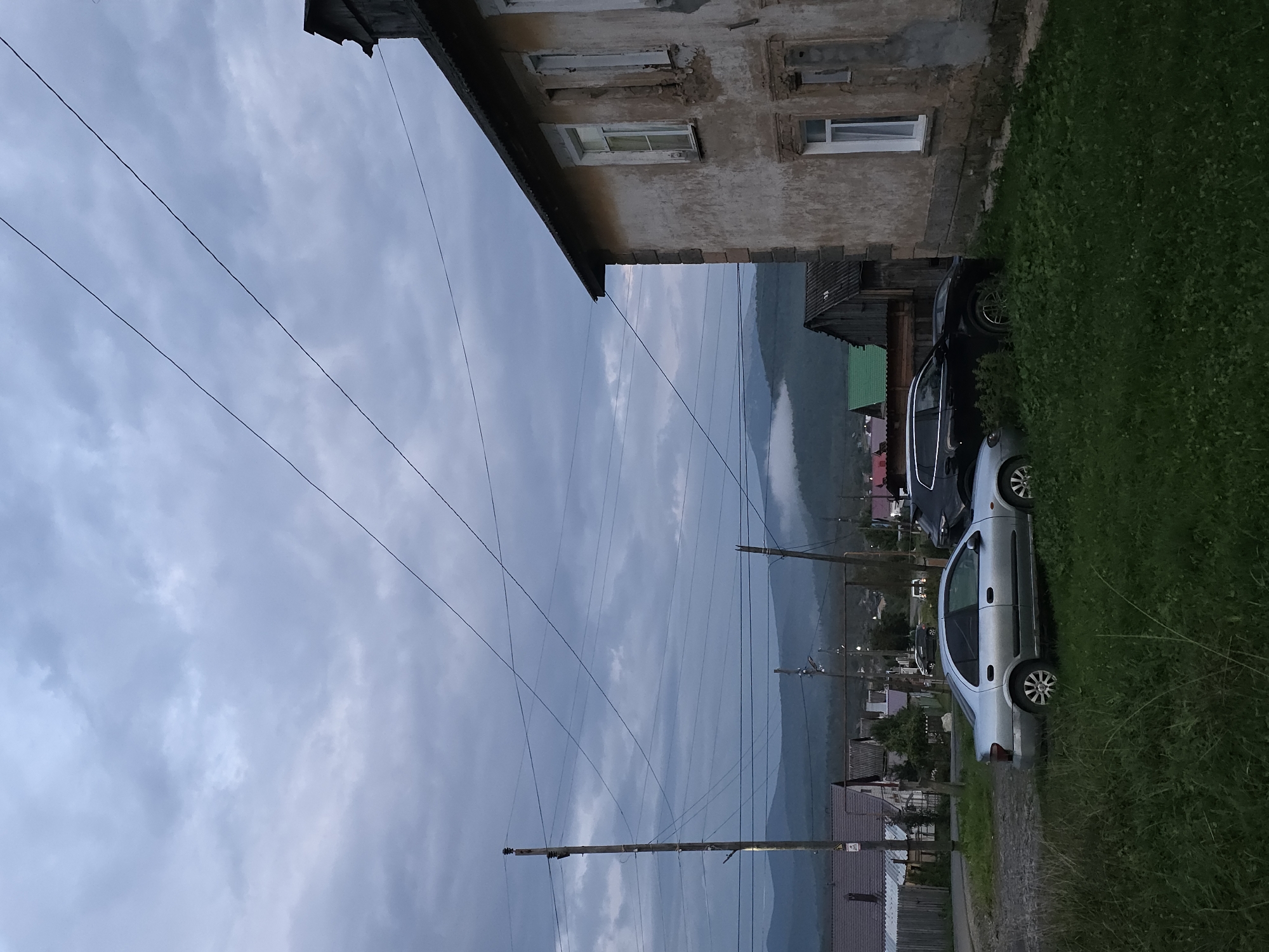 The image depicts a rural landscape with a cloudy sky. In the foreground, there is a grassy area and two cars parked on a gravel surface. To the right is a weathered, multi-story building with chipped plaster and visible stone work. In the background, a village with houses and a church is visible, nestled among rolling hills partially obscured by mist. Electrical poles and wires crisscross the scene. The location is identified as Magnitka, Russia. No people are visible in the image.