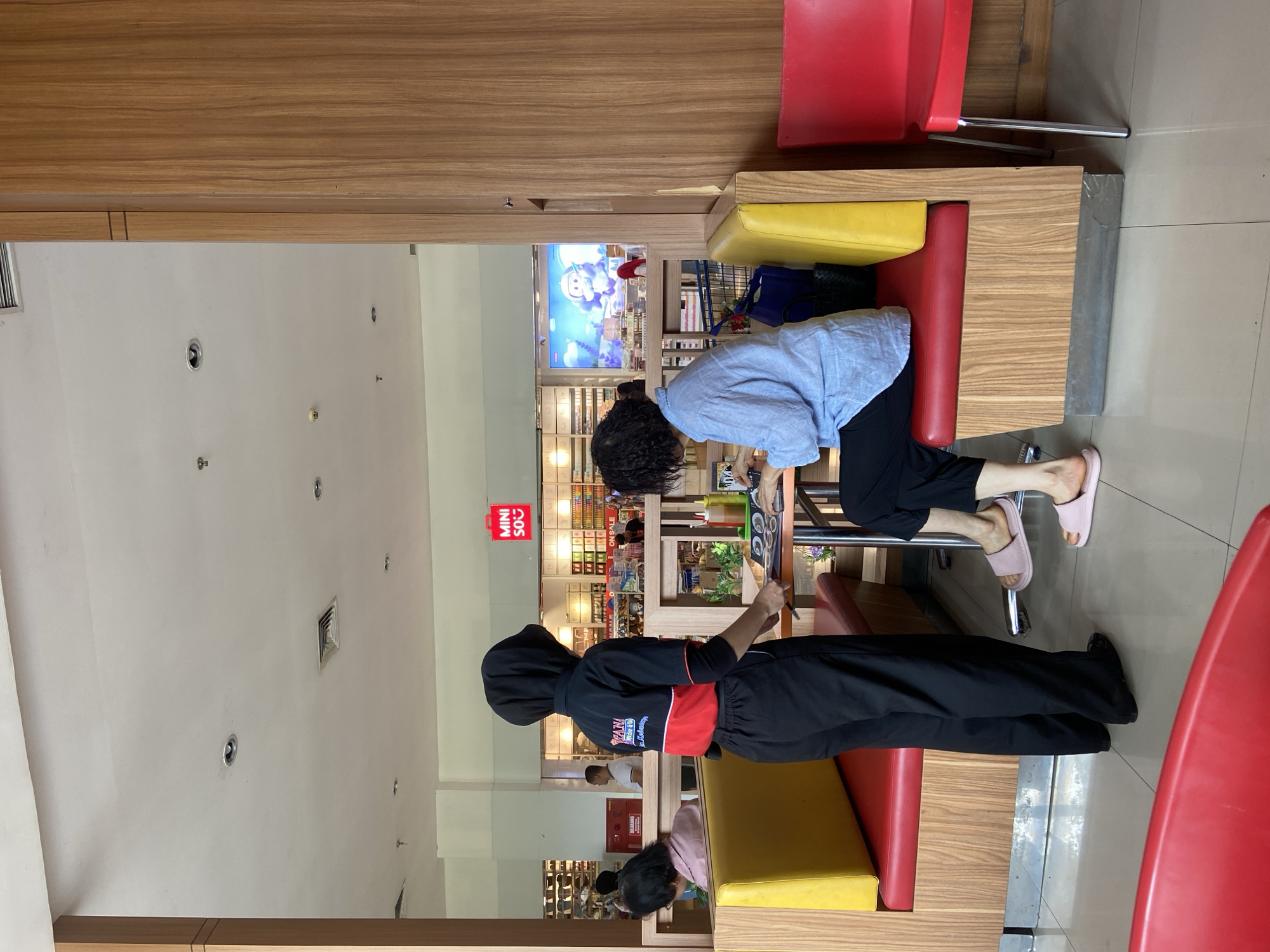 The image depicts the interior of a brightly lit, modern cafe in Kudus, Indonesia. A female server wearing a black headscarf, black pants, and a black and red uniform shirt stands to the left, holding a menu. She is interacting with a female customer who is seated at a table. The customer, dressed in a light blue shirt and black pants, is looking down at something on the table. The cafe features booth seating with red and yellow cushions, and wooden paneling. In the background, a store called "MINI SO" is visible, along with shelves stocked with merchandise. Another customer is seated at a booth in the background, to the left of the server. The floor is tiled and reflects the light.
