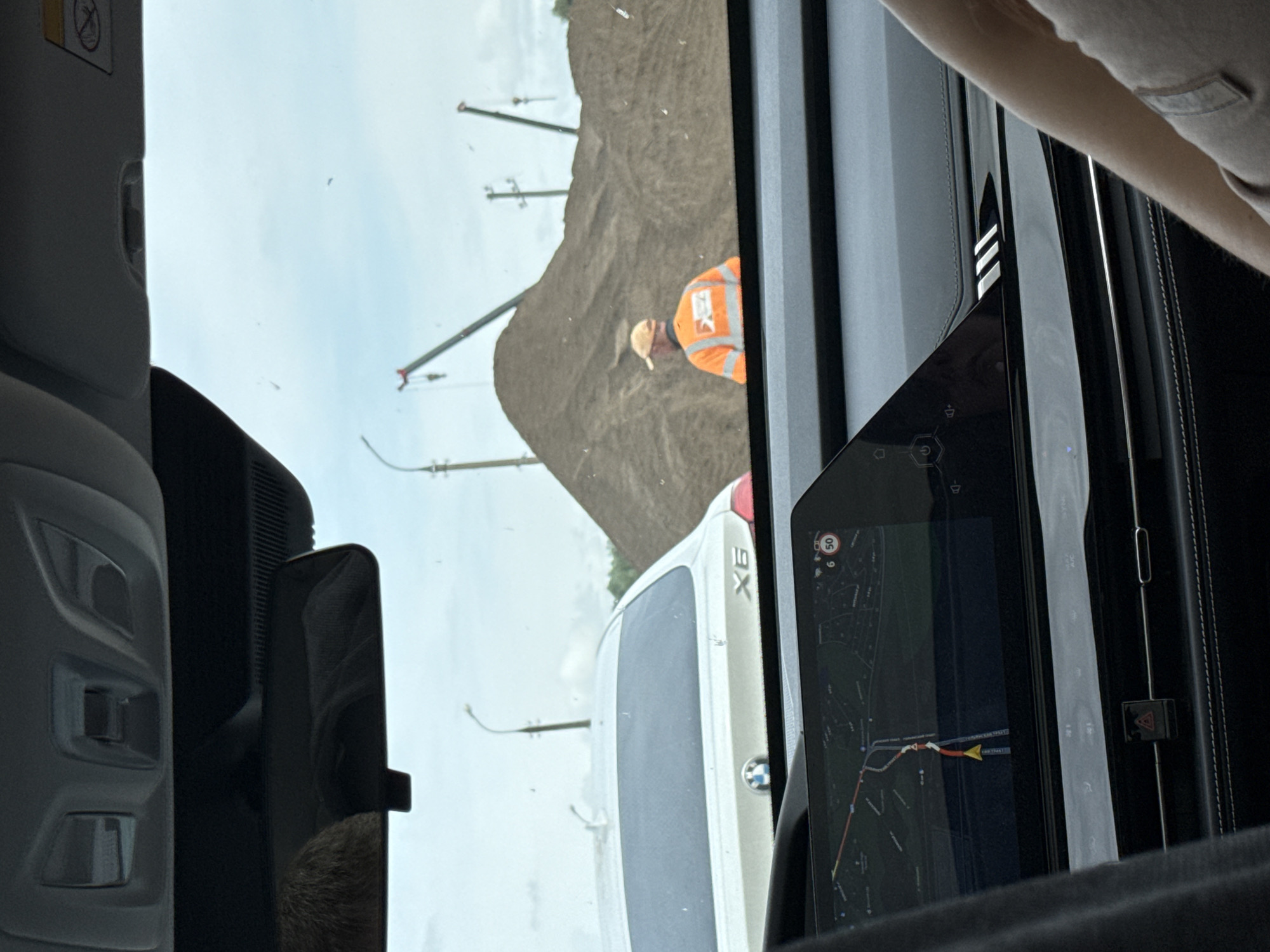The image, taken from inside a vehicle, looks out towards a roadside scene under an overcast daytime sky. The perspective is from the passenger side, with interior elements framing the view. On the far left, a grey car sun visor with a "no airbag" warning symbol is visible. Below it, part of the car's dark grey interior paneling is seen. On the right, a large digital display shows a navigation map with a route highlighted in red and yellow, indicating a speed limit of "50" in a circle and a number "6" (possibly indicating time or distance). A person's arm or shoulder is also visible on the far right.
Outside the vehicle, a construction or earthworks site is visible, dominated by a large brown dirt mound on the right. A construction worker wearing an orange high-visibility vest and a light-colored hard hat or cap stands near the dirt mound. Several tall, slender poles or gantries extend into the sky behind the mound and in the distance. Ahead of the viewing car, partially obscured by the car's interior, is the rear of a white BMW X6 SUV, with its "X6" badge and circular BMW logo clearly visible. The overall scene suggests travel through an area undergoing construction or industrial activity, possibly in Kabanikha, Russia, as indicated by the context.