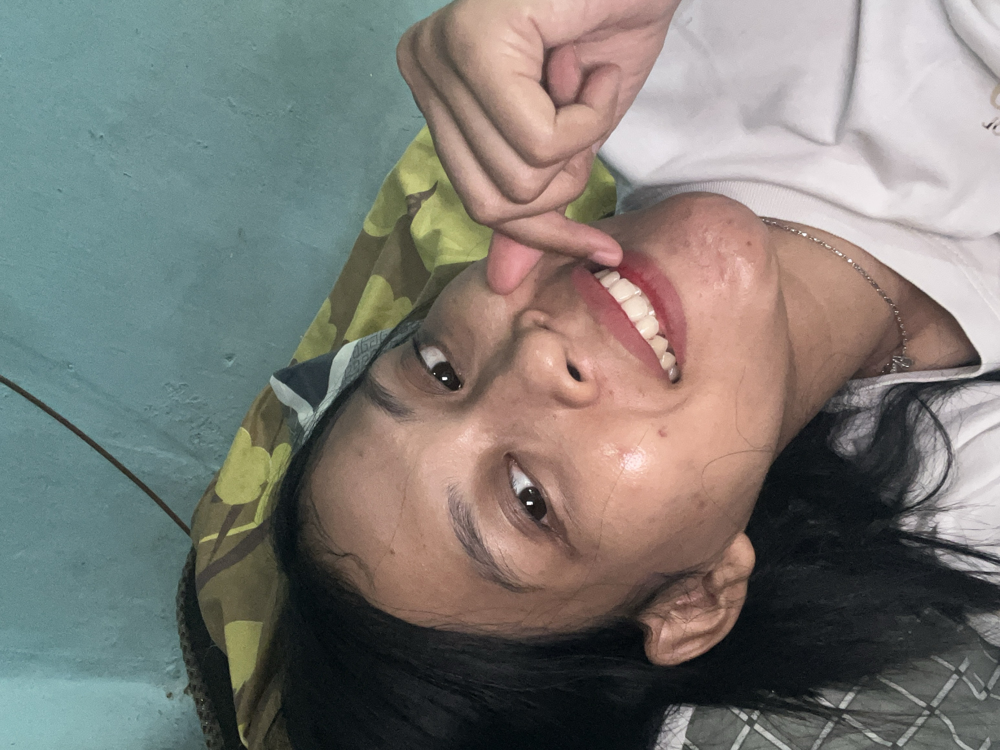 A woman lies on her side in bed, propping herself up on her elbow. She is wearing a white t-shirt and a silver necklace. Her dark hair is loose around her face and shoulders. She is smiling broadly, her lips parted to reveal white teeth. Her right hand is raised, with her index finger and thumb touching her cheek. The background shows a blue wall and a pillow with a yellow and green pattern. The overall impression is of a relaxed, informal moment captured in a bedroom in Kecamatan Pati, Indonesia.
