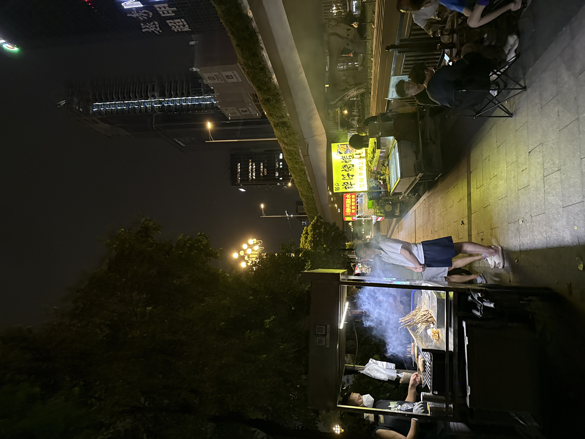 In a dimly lit urban setting, likely in Huangpu District, China, a street food stall is the focal point. The time appears to be night, indicated by the dark sky and artificial lighting. The stall is set up on a paved sidewalk next to a grassy median. A building with numerous illuminated windows is visible in the background, suggesting a commercial or residential area.
Several people are present. Two individuals are seated on chairs near the food stall, engrossed in their phones. Another person, wearing a white shirt and dark shorts, stands near the stall, seemingly waiting or observing. Further down the sidewalk, another person is seated, wearing glasses and a dark shirt. A vendor is actively preparing food at the stall, with smoke rising from the cooking area. Two individuals are also seen standing near the food stall.
The food stall itself is a prominent object, equipped with a display case for food items and a cooking area. The text visible on the stall and surrounding signage includes "老曾螺蛳粉", "牌仔燒烤", and "湿辣牛肉2". These indicate that the stall sells螺蛳粉 (Luosifen, a type of noodle soup) and烧烤 (shaokao, grilled skewers), specifically mentioning spicy beef skewers. The overall atmosphere suggests a typical evening street food scene, with people gathering to eat and socialize. The weather appears clear.